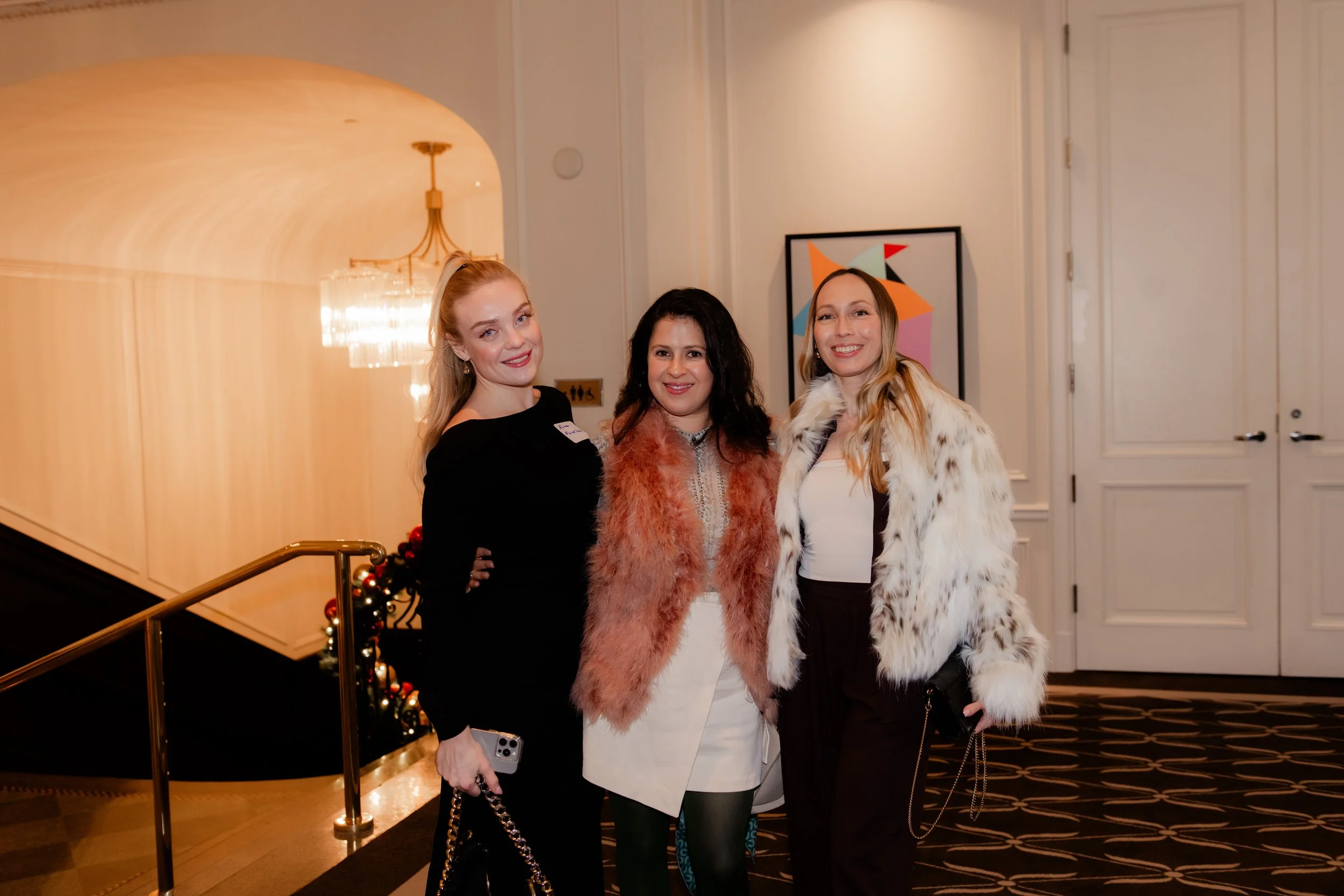 Three women dressed elegantly standing together in an indoor setting, smiling at the camera.
