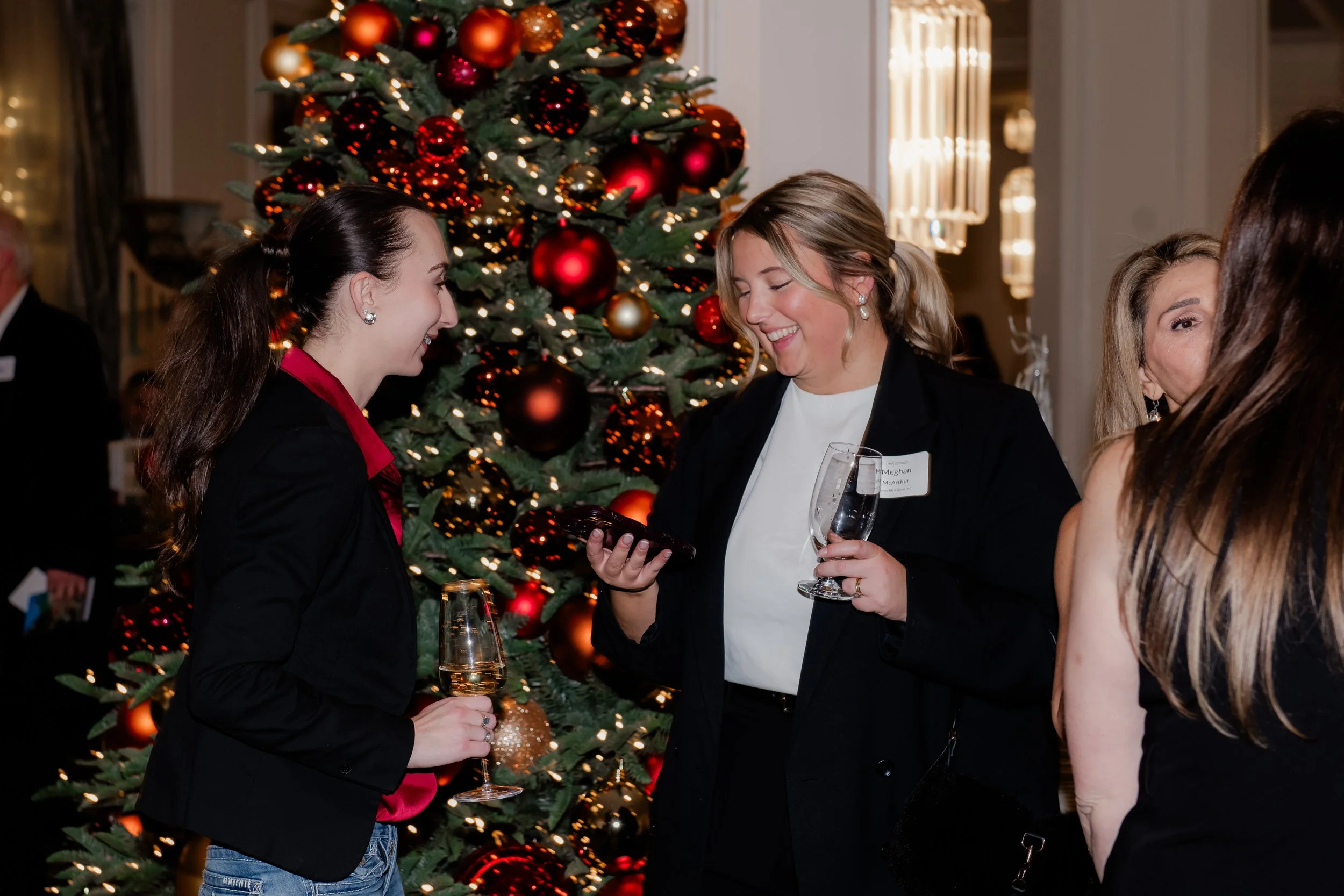 Women at a Christmas party chatting by a decorated Christmas tree, holding glasses of wine.