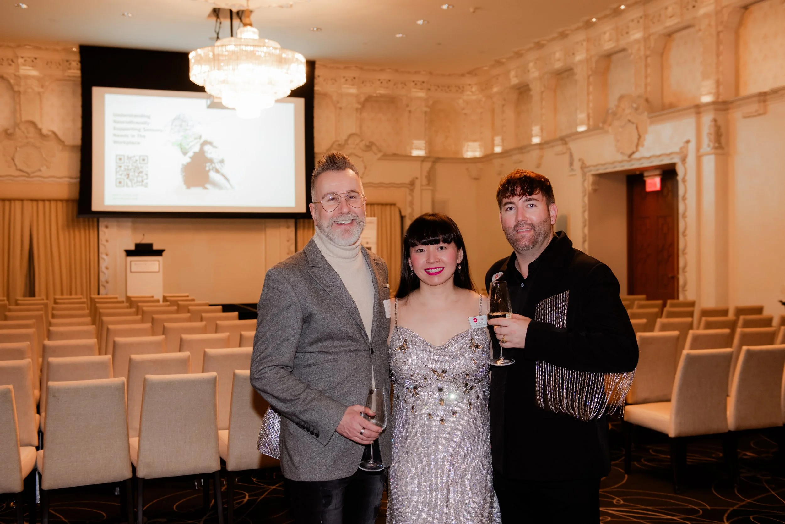 Three people at a formal event holding drinks in a decorated hall with a chandelier, facing the camera.