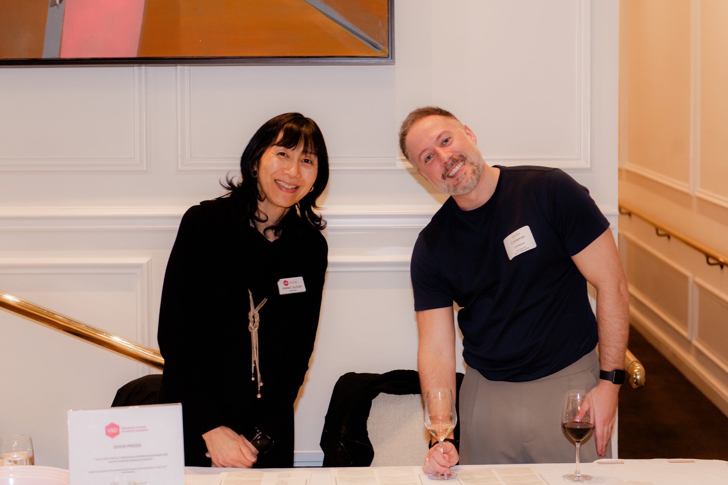 Two people at a formal event, smiling and holding glasses of wine, standing behind a table with a sign and glasses, in a well-lit room with white paneled walls and artwork.