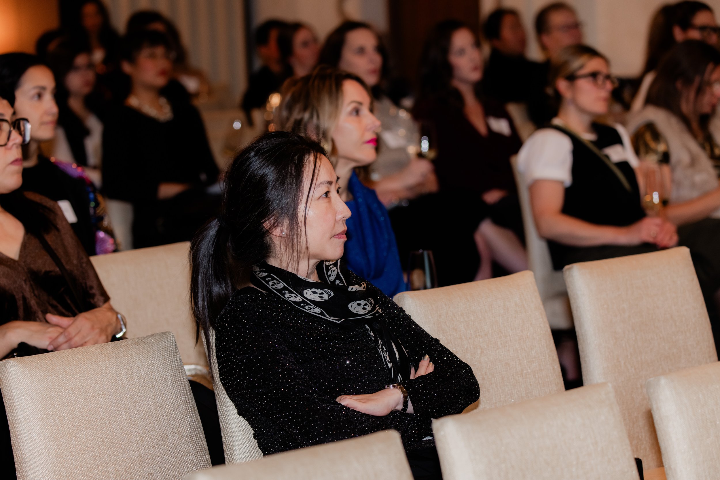 A group of women seated in an auditorium, attentively listening, some holding drinks.