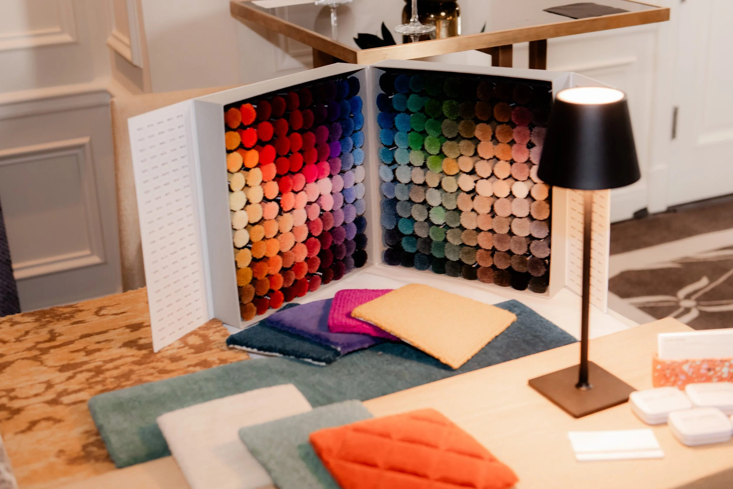 Color samples, fabric swatches, and a large color palette display on a table, with a black desk lamp and some other small items nearby.