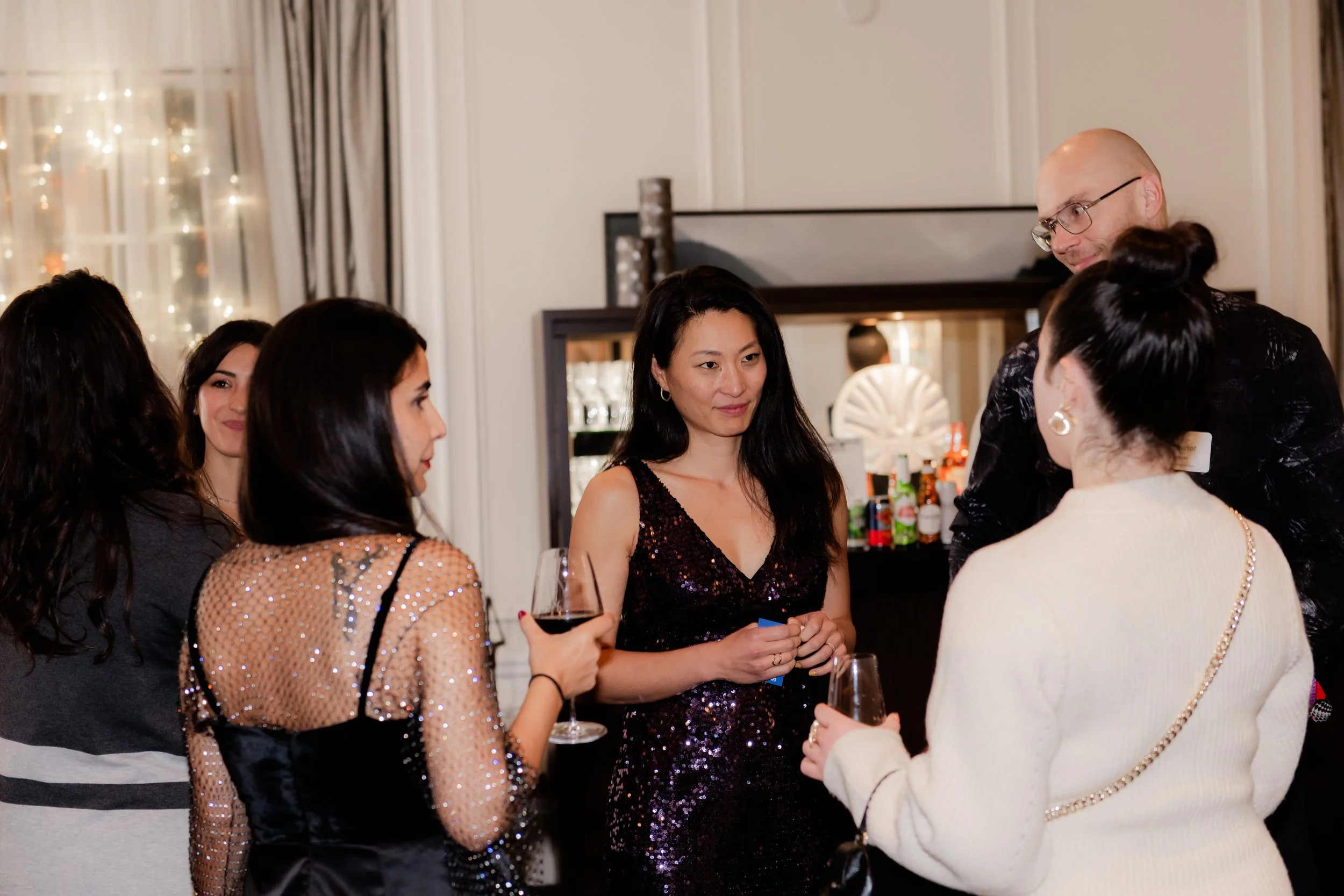 Group of people at a party, some holding glasses of wine, engaging in conversation in a well-lit room with a bar in the background.