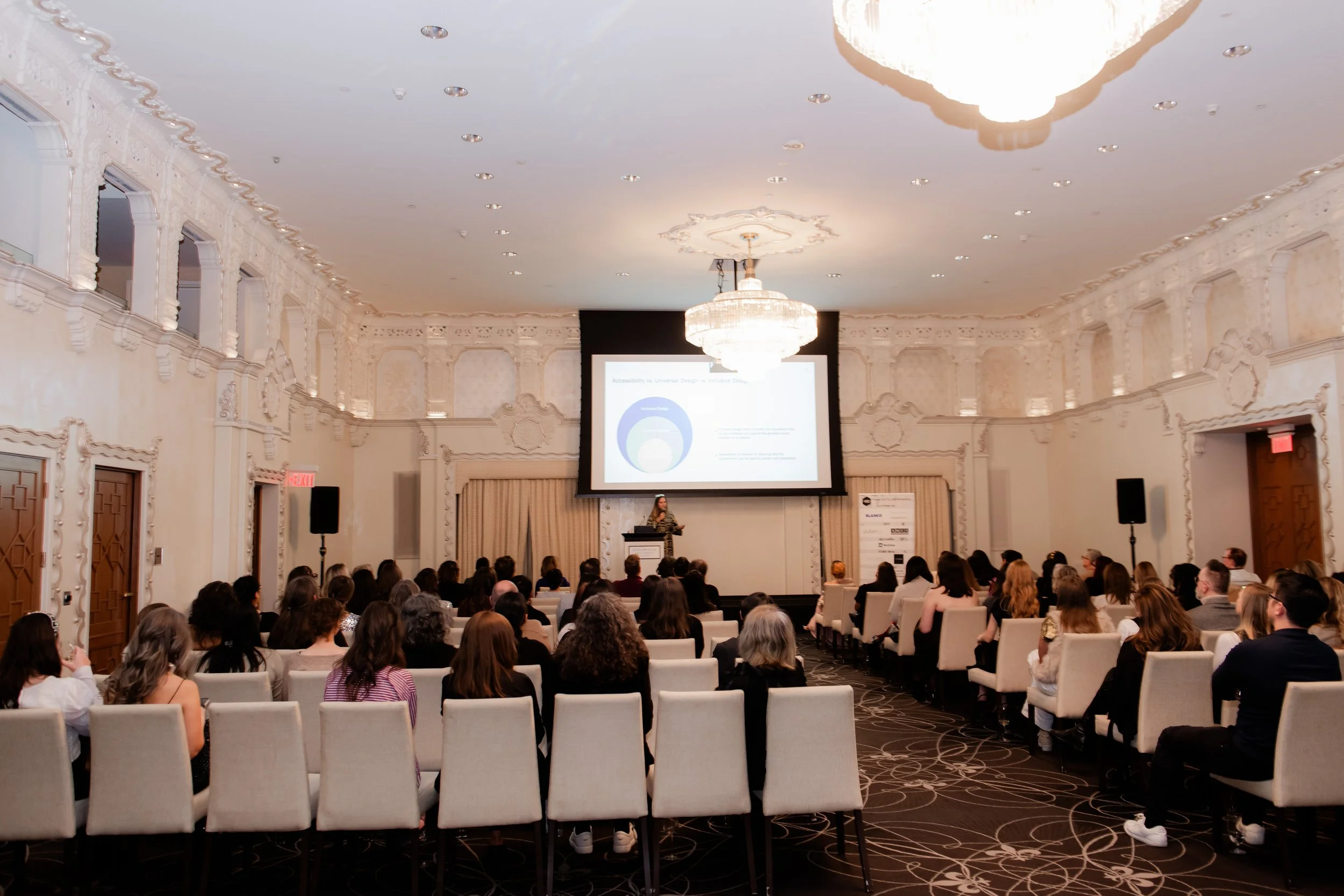 A professional conference or seminar taking place in an elegant room with a large chandelier, a speaker on stage presenting a slide, and an audience seated facing the stage.