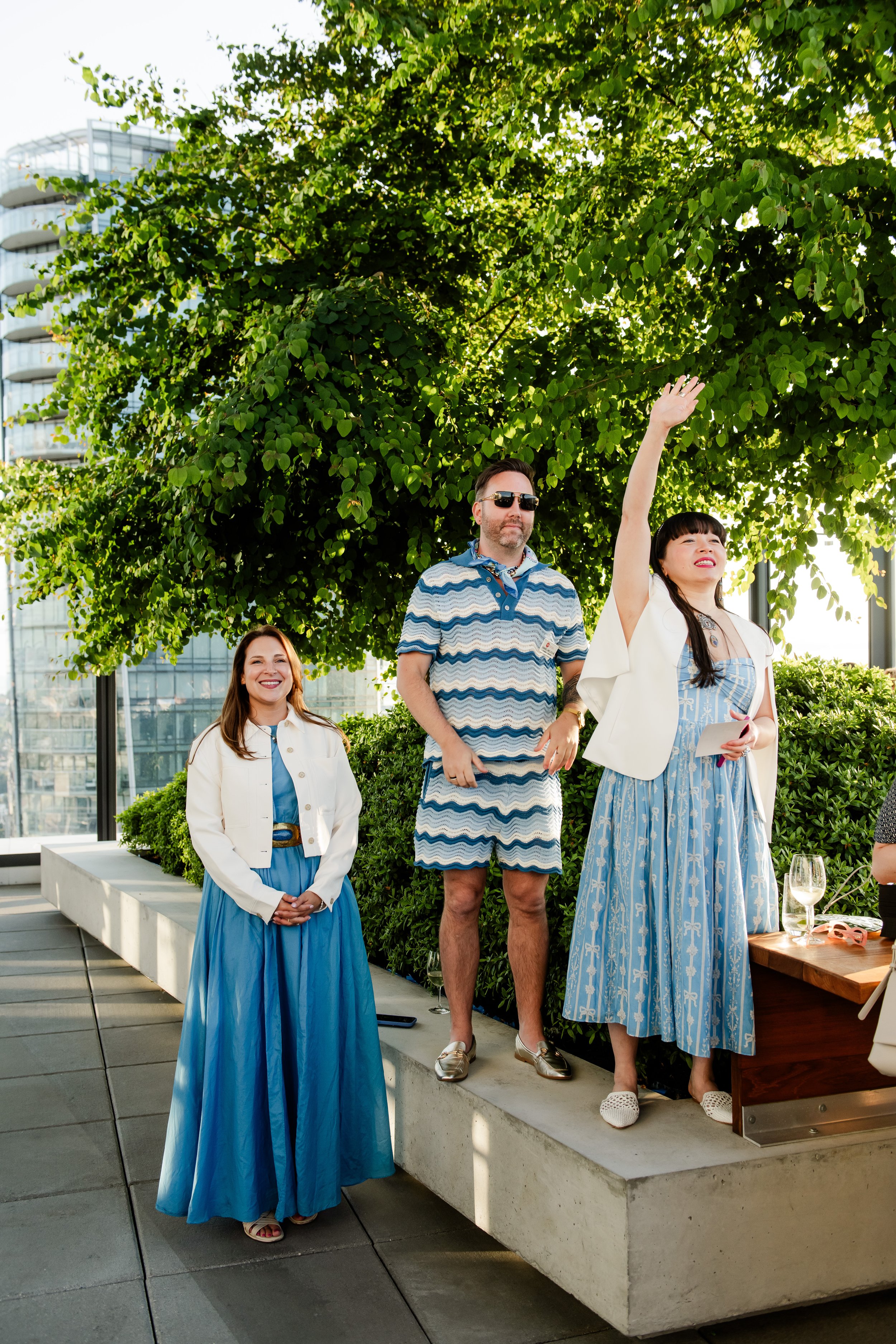 People dressed in summer outfits standing on a rooftop terrace, with greenery and modern building in the background, celebrating or socializing.