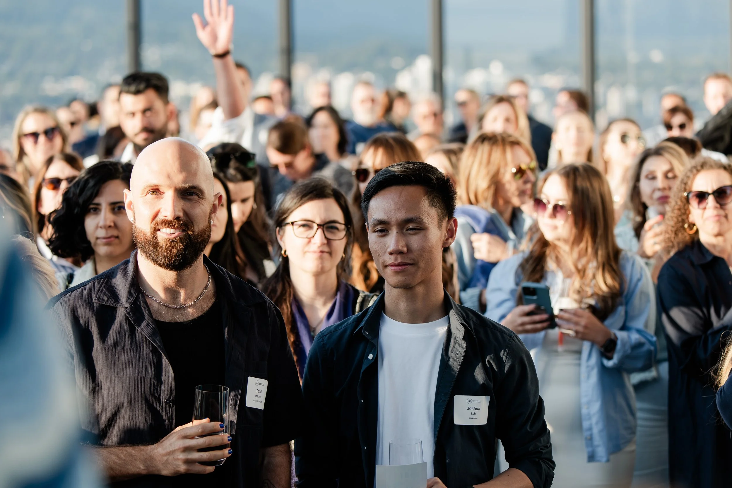 A large group of people at a social event or gathering, with two men in the foreground holding glasses, one smiling and the other serious, among a diverse crowd with some wearing sunglasses, in a well-lit space with large windows showing a cityscape 