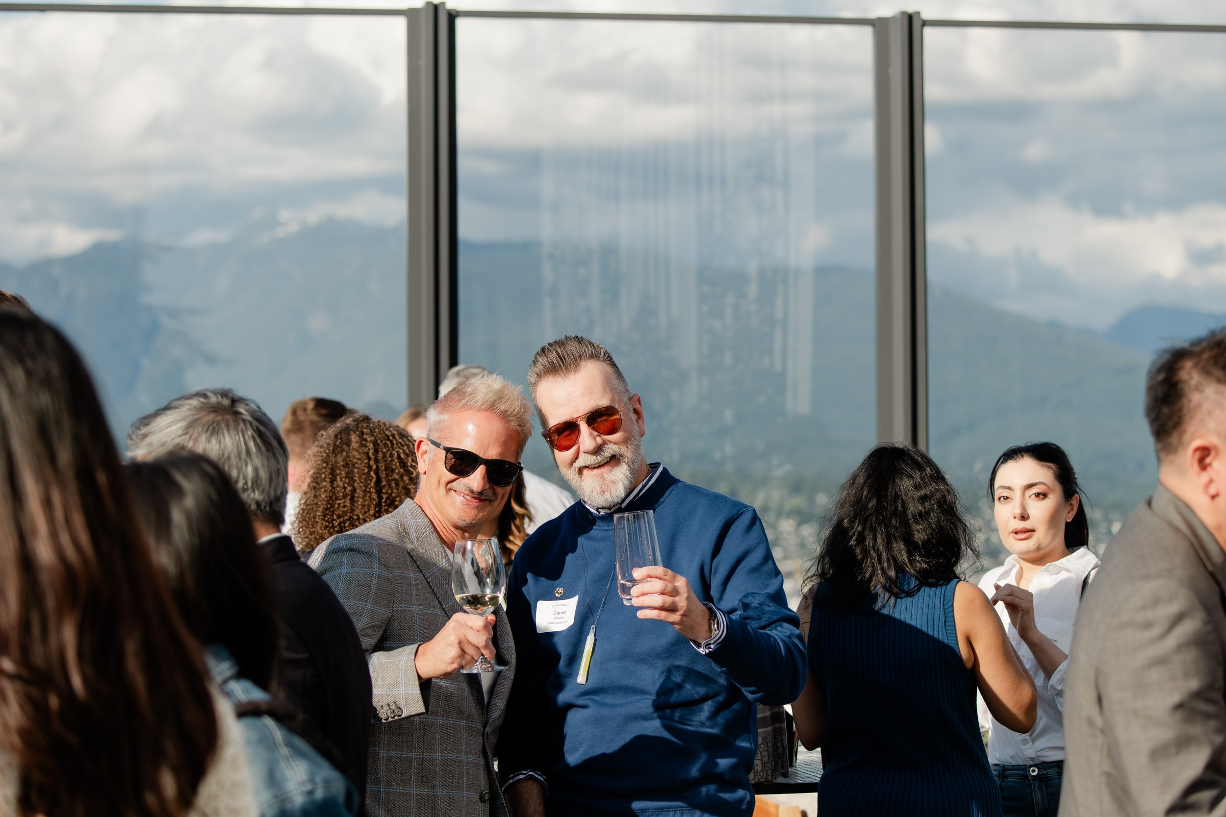 Two men smiling and holding wine glasses at a social gathering in a high-rise venue with large windows and mountain views