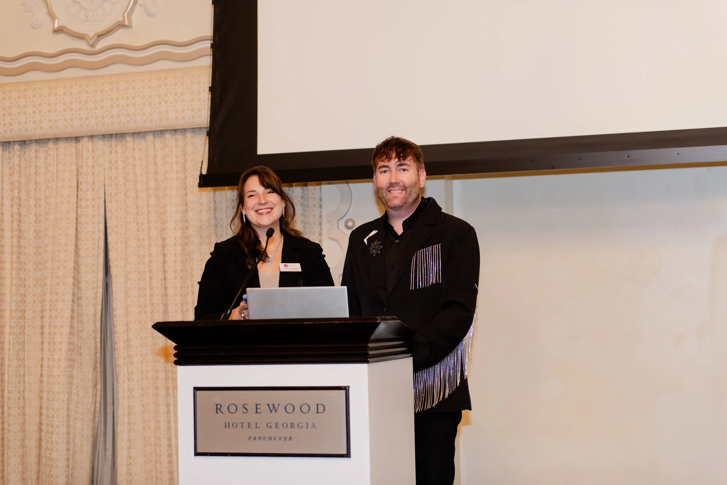 Two people standing behind a podium with a microphone at an event at Rosewood Hotel Georgia in Vancouver.