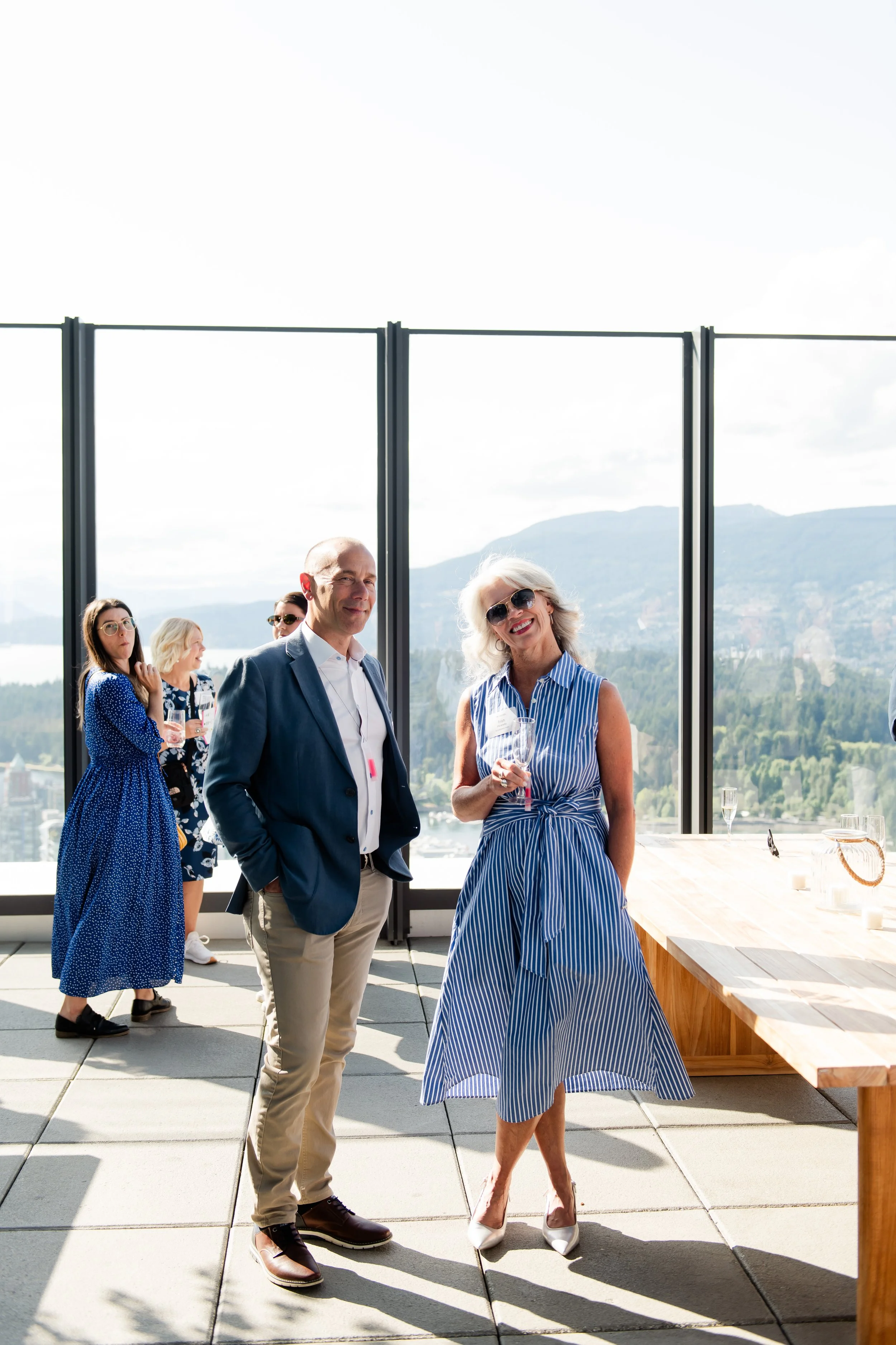 Group of people at outdoor rooftop event, with city skyline and mountains in background, some holding glasses of champagne.