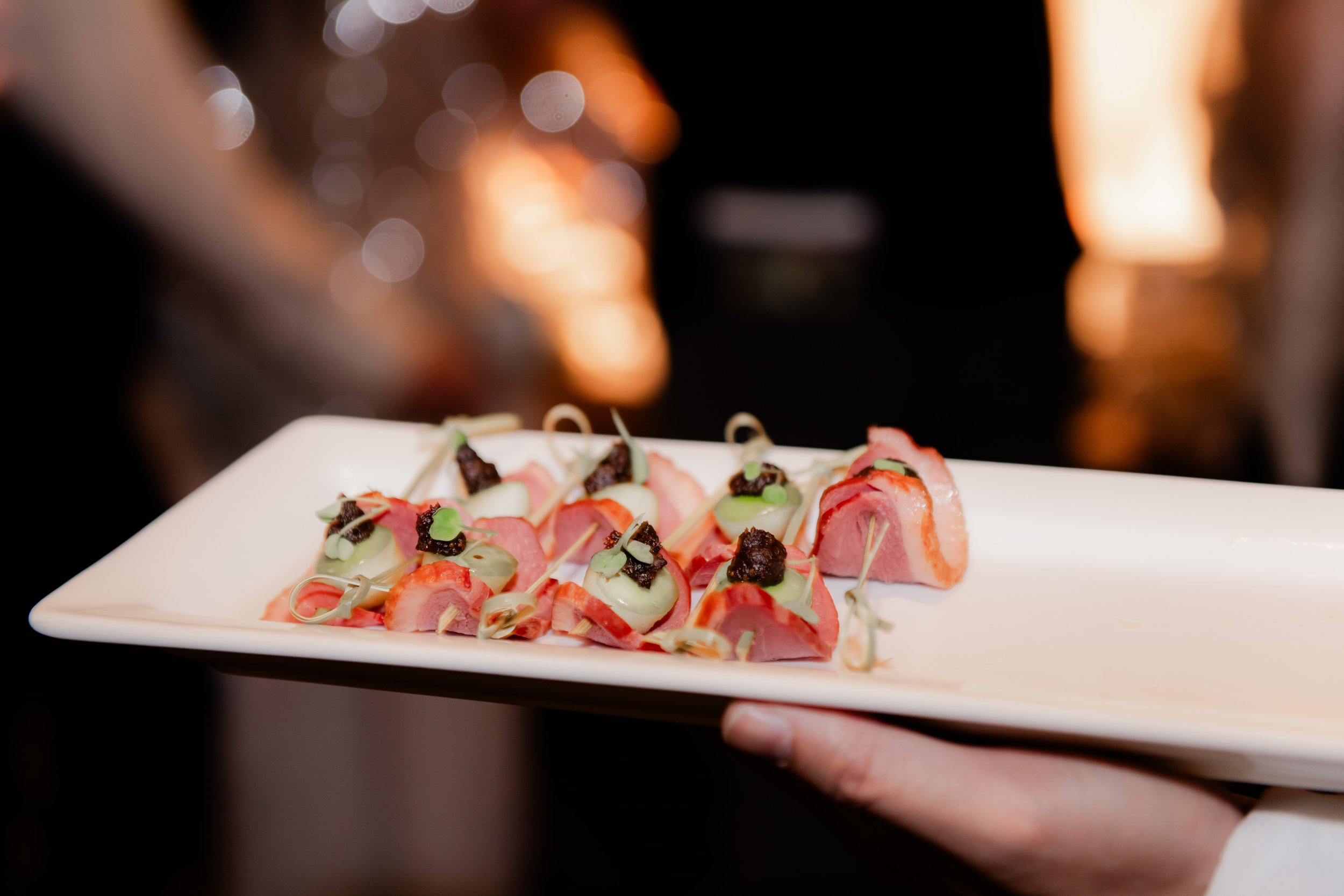 A person holding a white rectangular plate with small gourmet bites of pink meat, green sauce, and garnishes, served at a formal event.