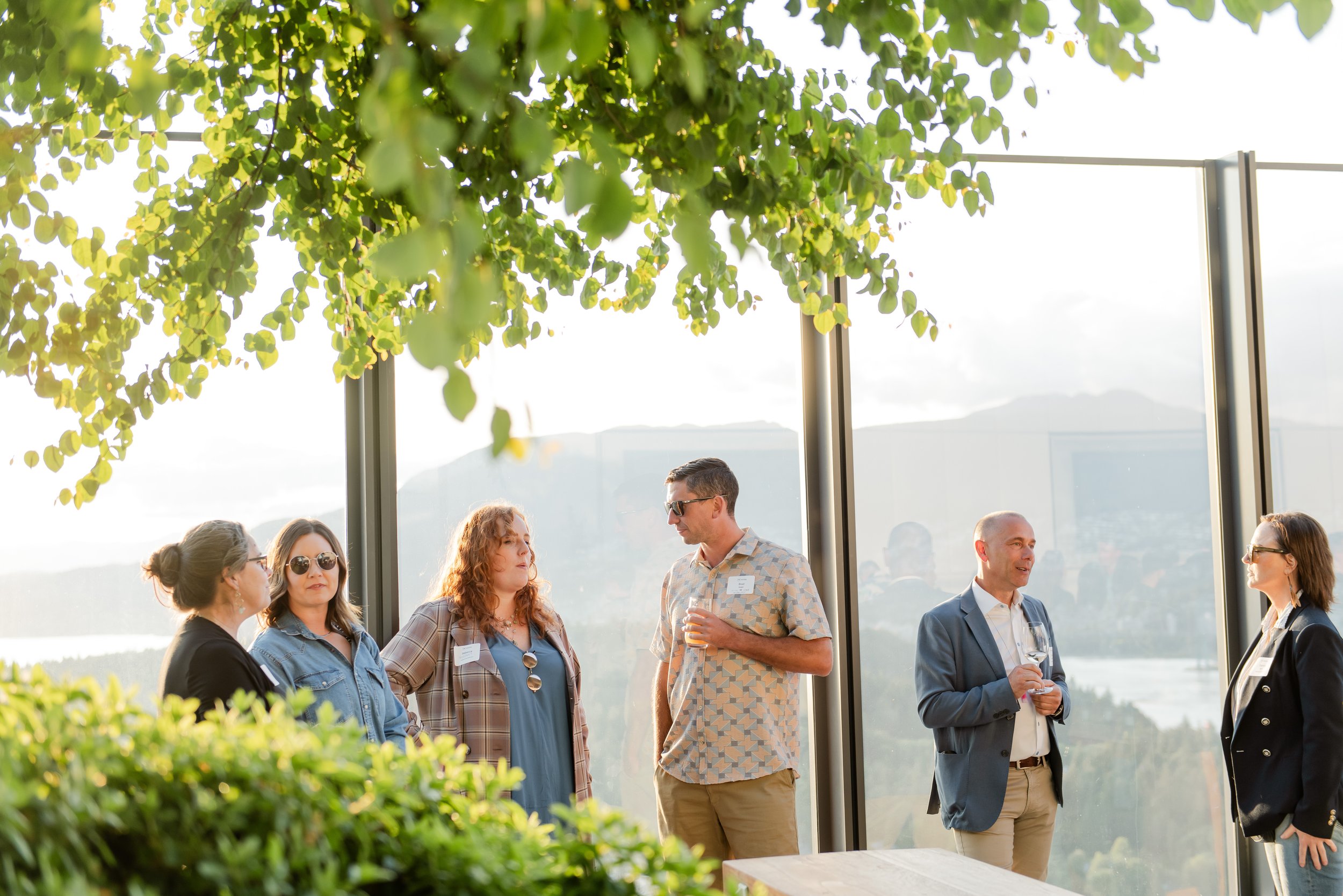 Group of six people socializing during an outdoor event, standing near a glass wall with a scenic mountain view, under a leafy tree.