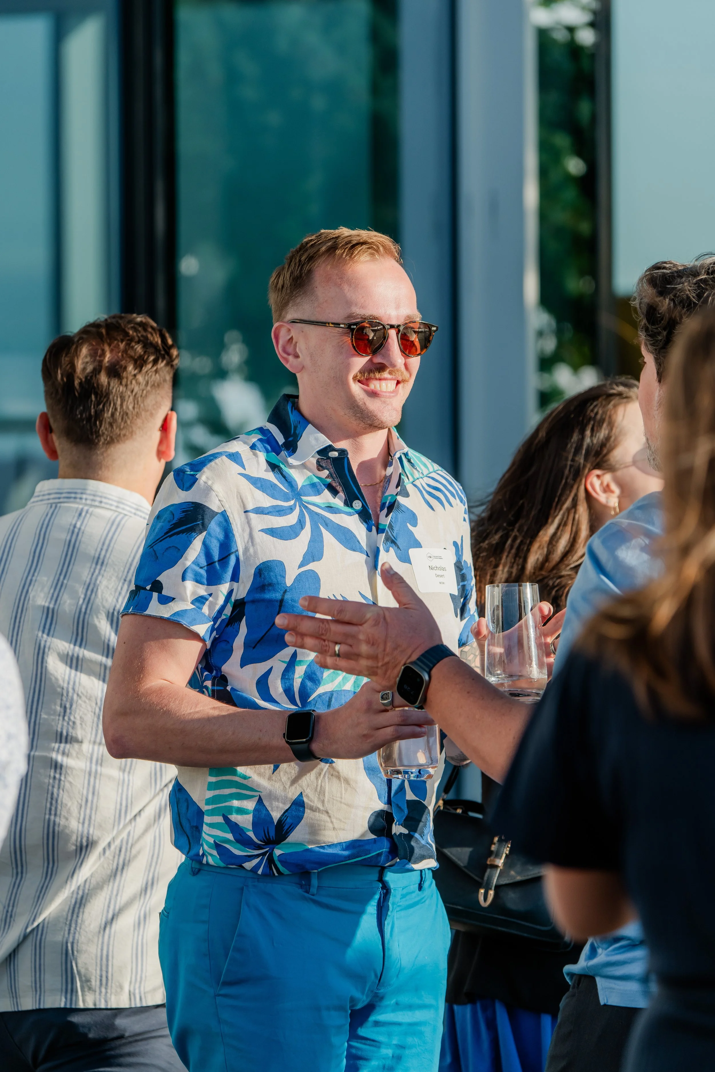 A man in a blue floral shirt and blue shorts wearing sunglasses, holding a glass, smiling, and engaging in conversation at a social event indoors.