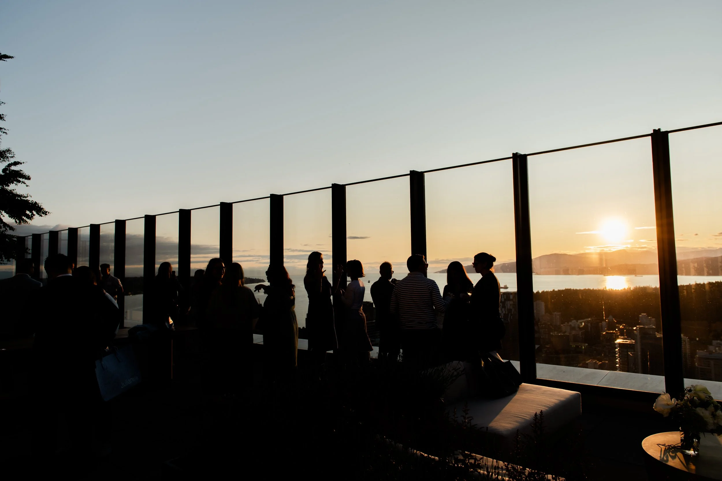 People gathered on a rooftop terrace during sunset, silhouetted against the view of a city skyline, river, and mountains in the distance.