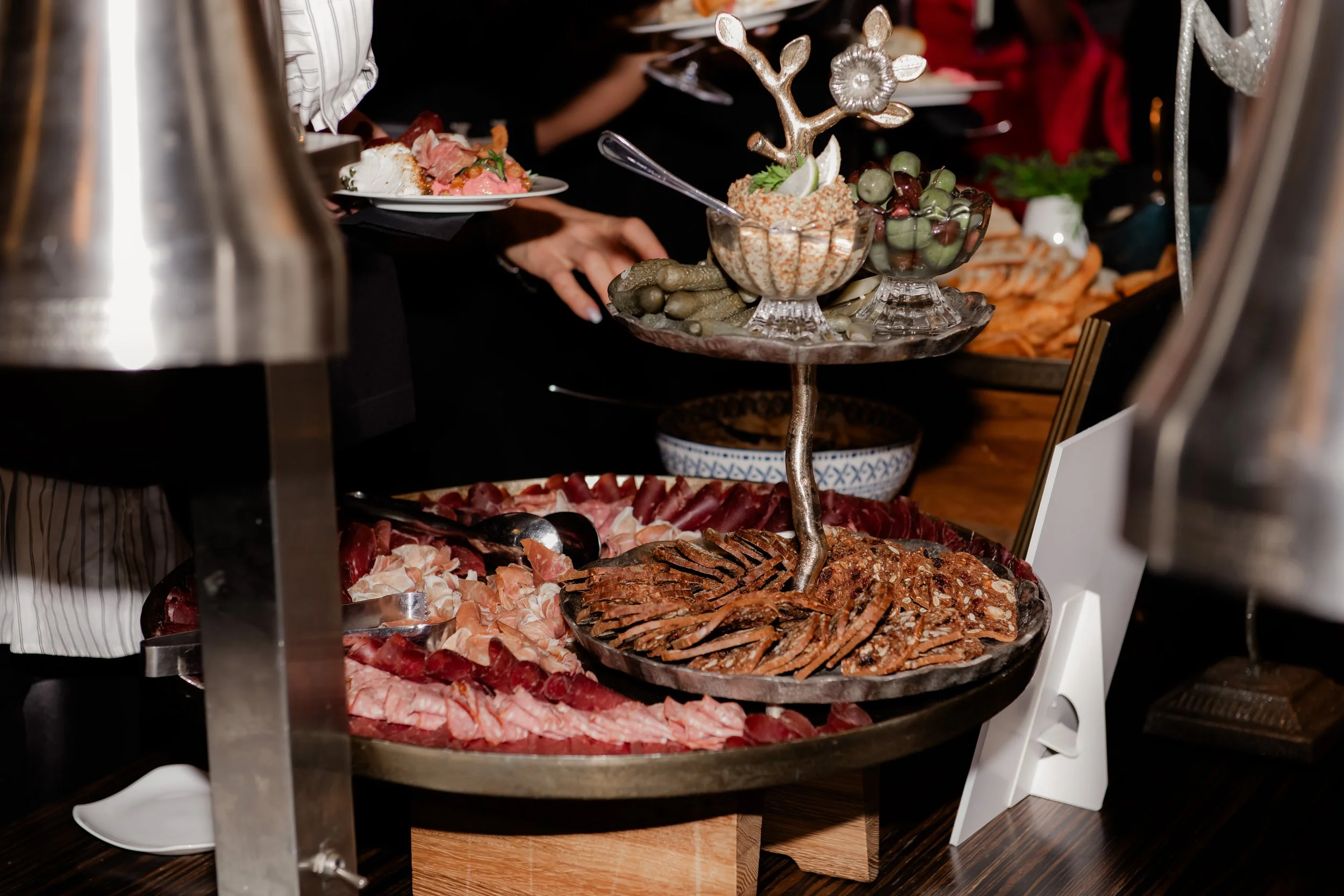 A buffet with sliced meats, a three-tiered serving tray with bowls of olives and dip, and salad on a plate in the background.