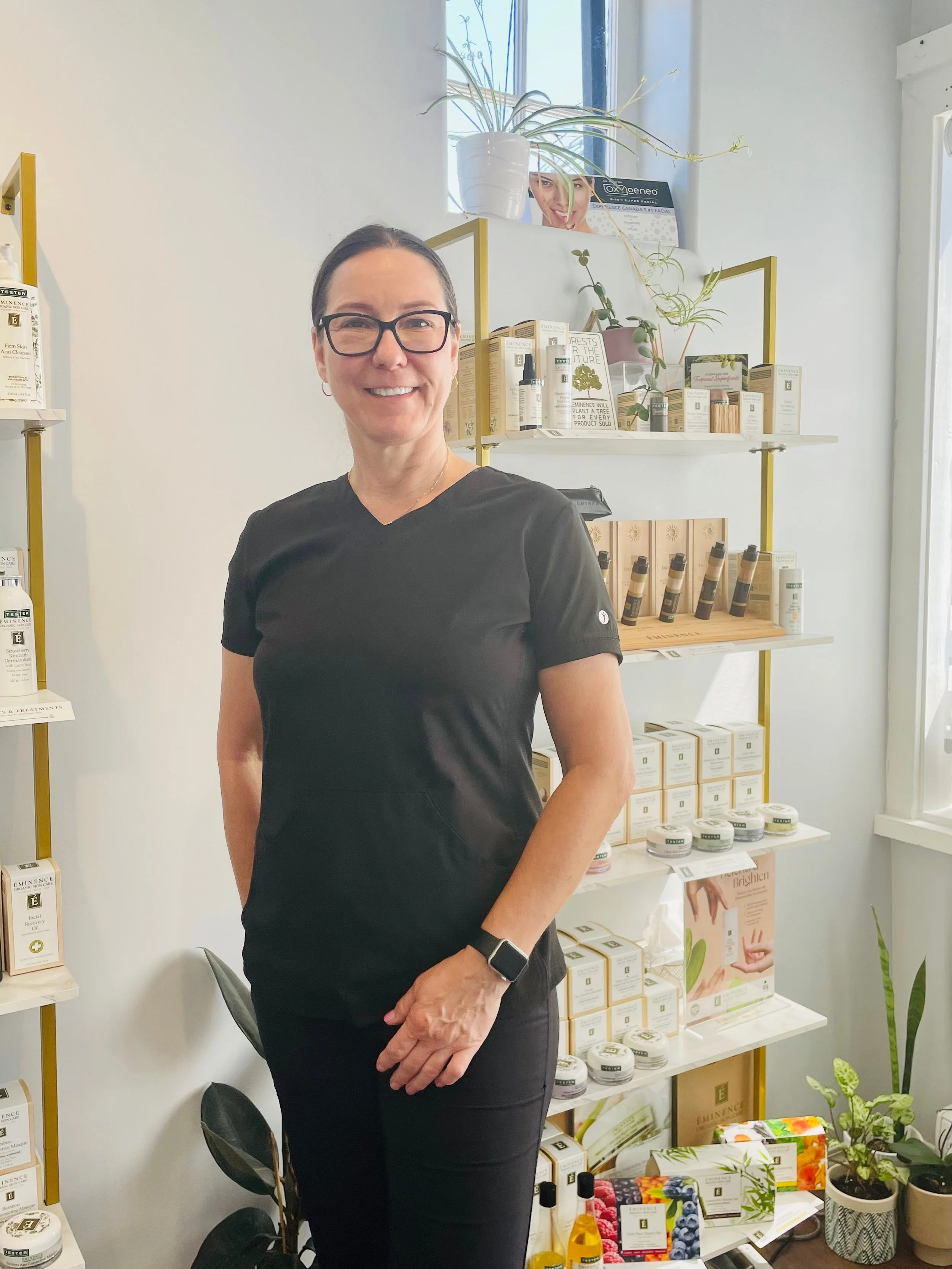 A woman wearing glasses and black scrubs standing in front of shelves stocked with skincare products in a bright room with a window.