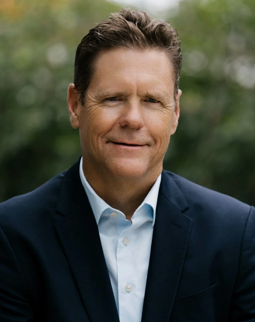 A middle-aged man with brown hair, wearing a dark blazer over a light blue collared shirt, smiling outdoors with a blurred green background.