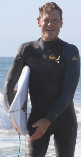 Man in a wetsuit holding a surfboard on a beach with the ocean behind him.