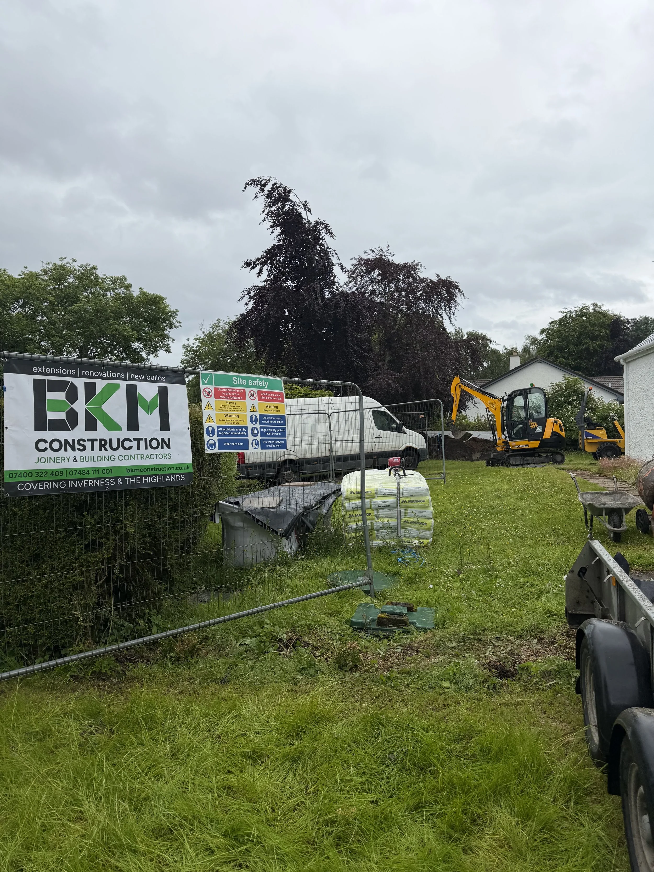 Construction site with fencing, construction vehicles, a sign for BKM Construction, and a grassy field with trees and houses in the background.