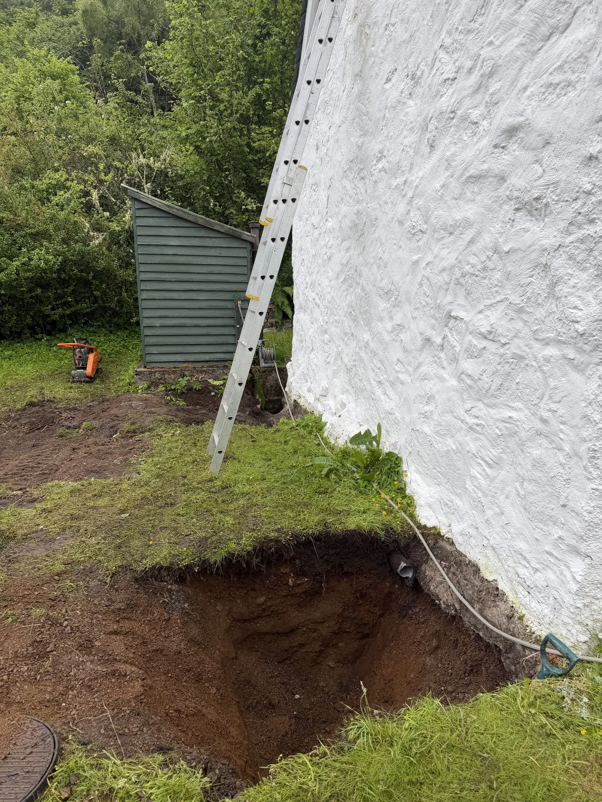 A large hole in the ground next to the side of a white stucco building, with a metal ladder leaning against the building and a small shed in the background. The area around the hole is grassy, with some plants and outdoor equipment nearby.