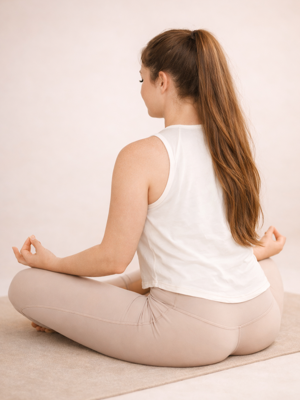 Woman seated cross‑legged on a beige mat against a soft neutral background.
