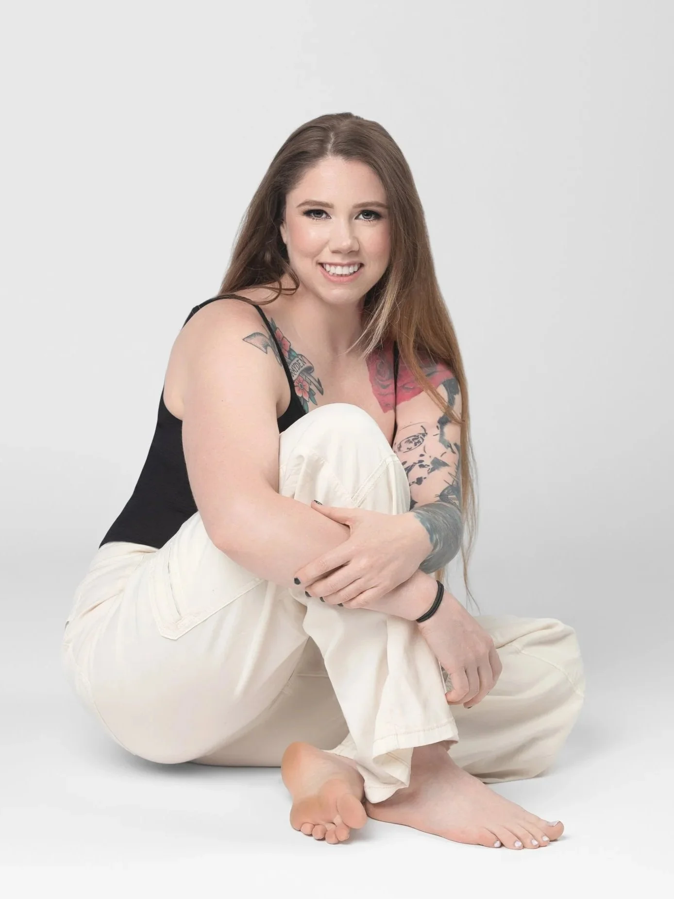 A young woman with long brown hair, tattoos on her arms and shoulder, wearing a black tank top and cream pants, sitting barefoot on a plain light gray background, smiling and looking at the camera.