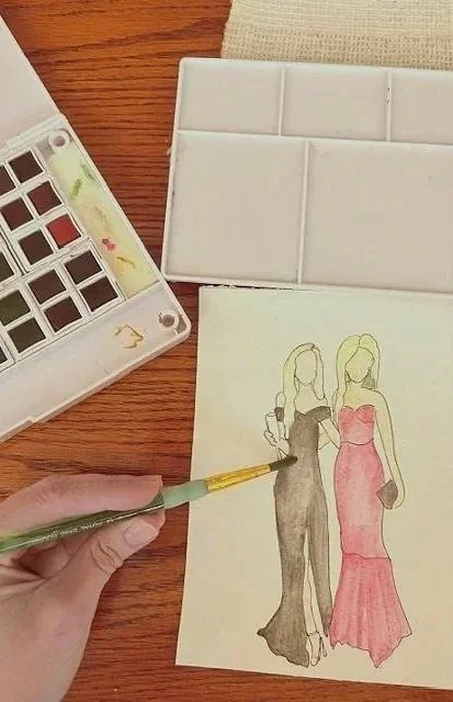 A hand holding a paintbrush painting two women in elegant dresses on paper. One woman is wearing a black dress, and the other is in a pink dress. Art supplies, including watercolor paints and a watercolor palette, are visible on the wooden table.