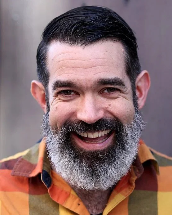 A man with a thick salt-and-pepper beard smiling outdoors, wearing a colorful plaid shirt.