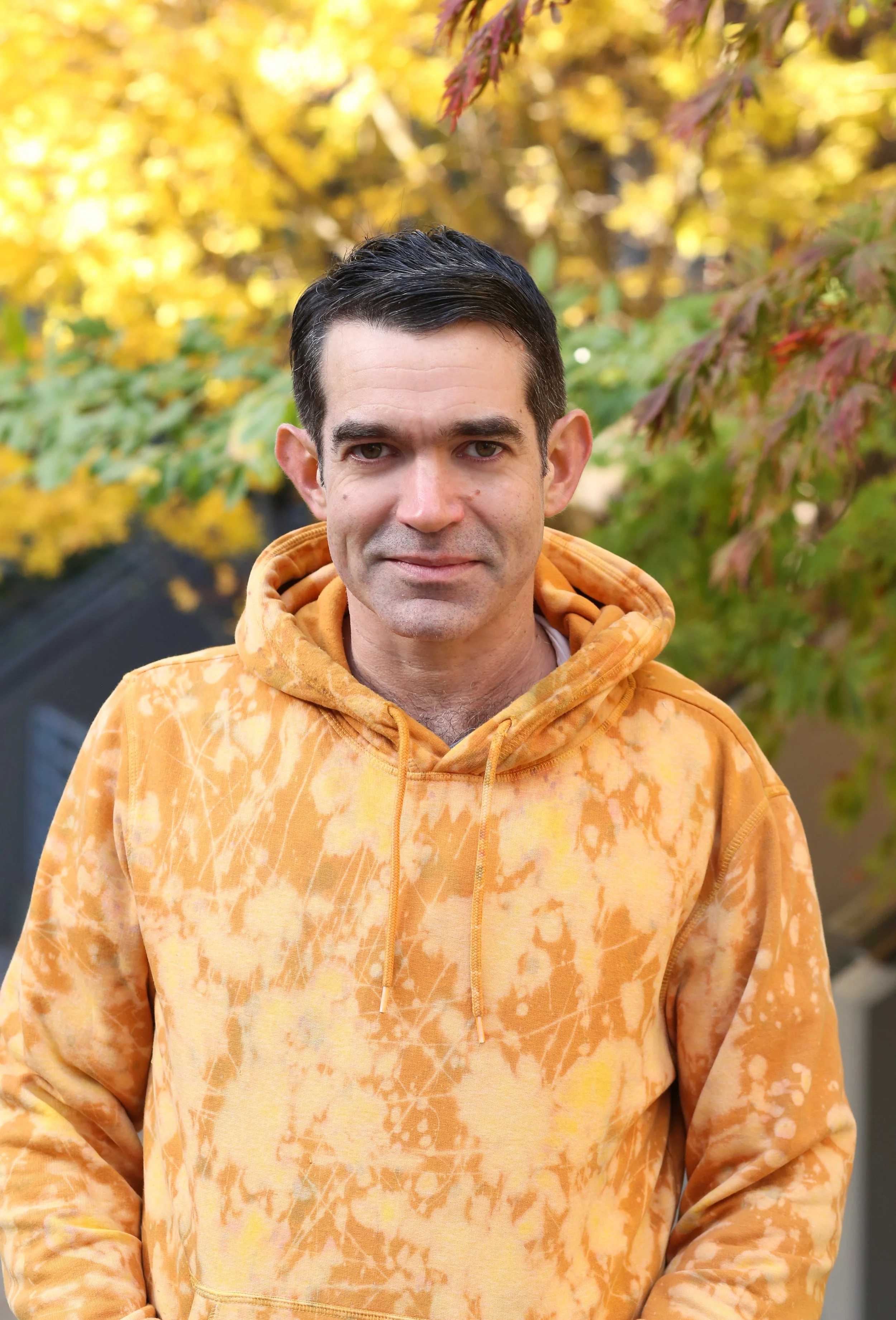A man standing outdoors in front of trees with autumn-colored leaves, wearing an orange tie-dye hoodie.