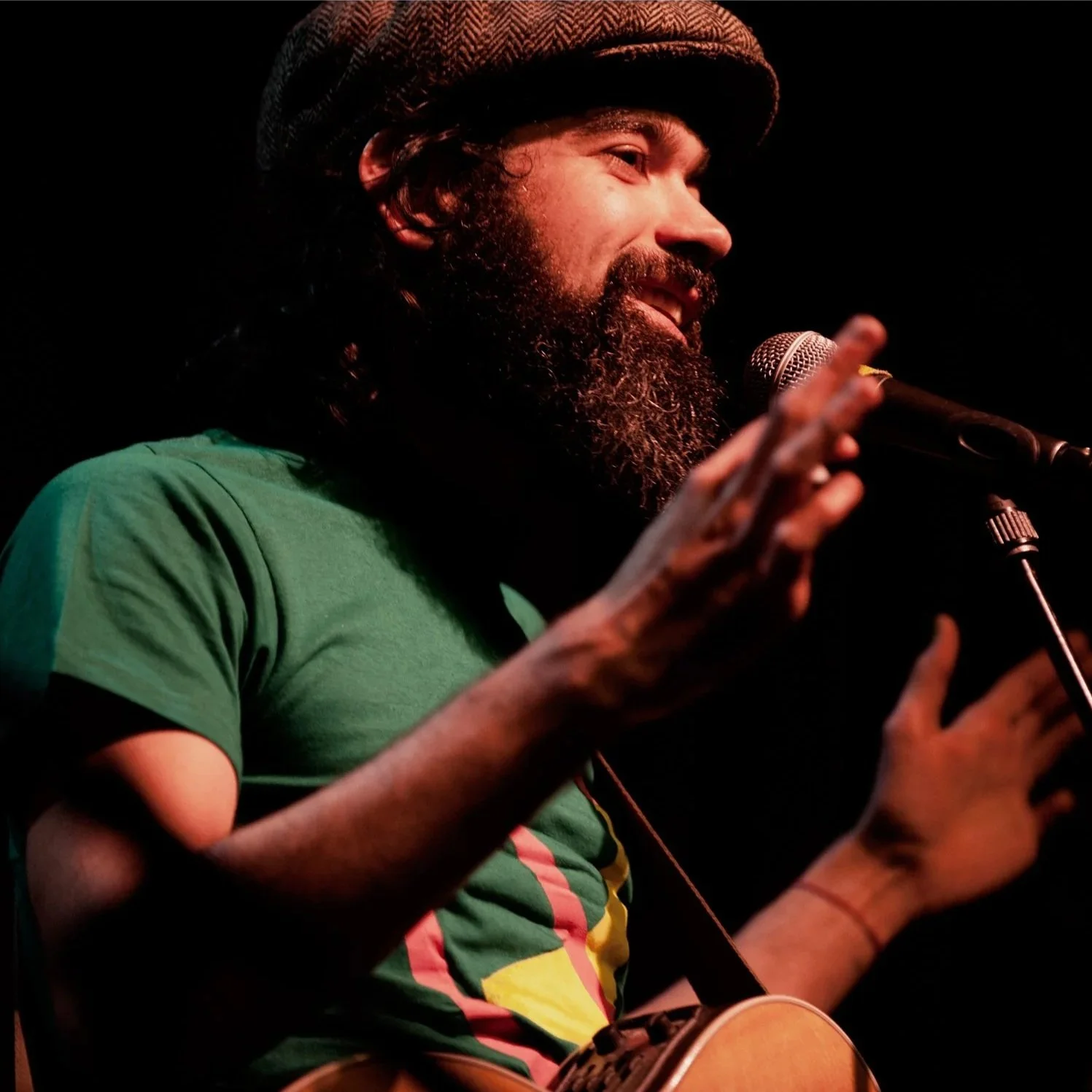 A bearded man wearing a brown cap and a green T-shirt is smiling and speaking or singing into a microphone while playing an acoustic guitar.