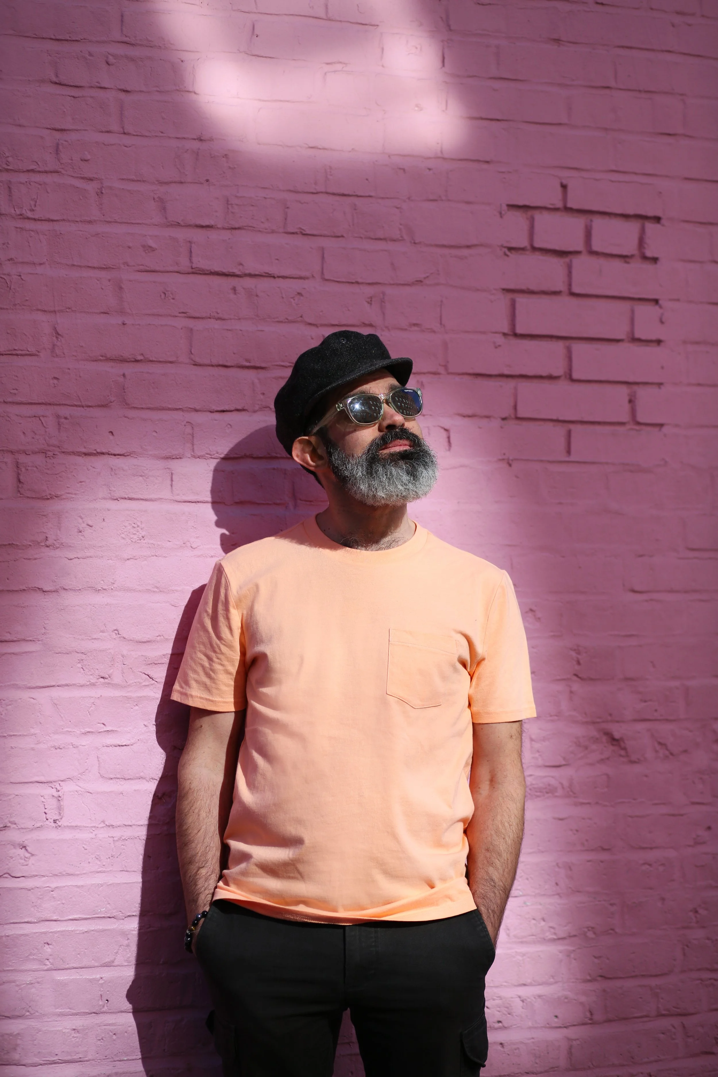 A man with a gray beard wearing a black hat, round reflective sunglasses, a light peach t-shirt, and black pants, standing against a pink brick wall.