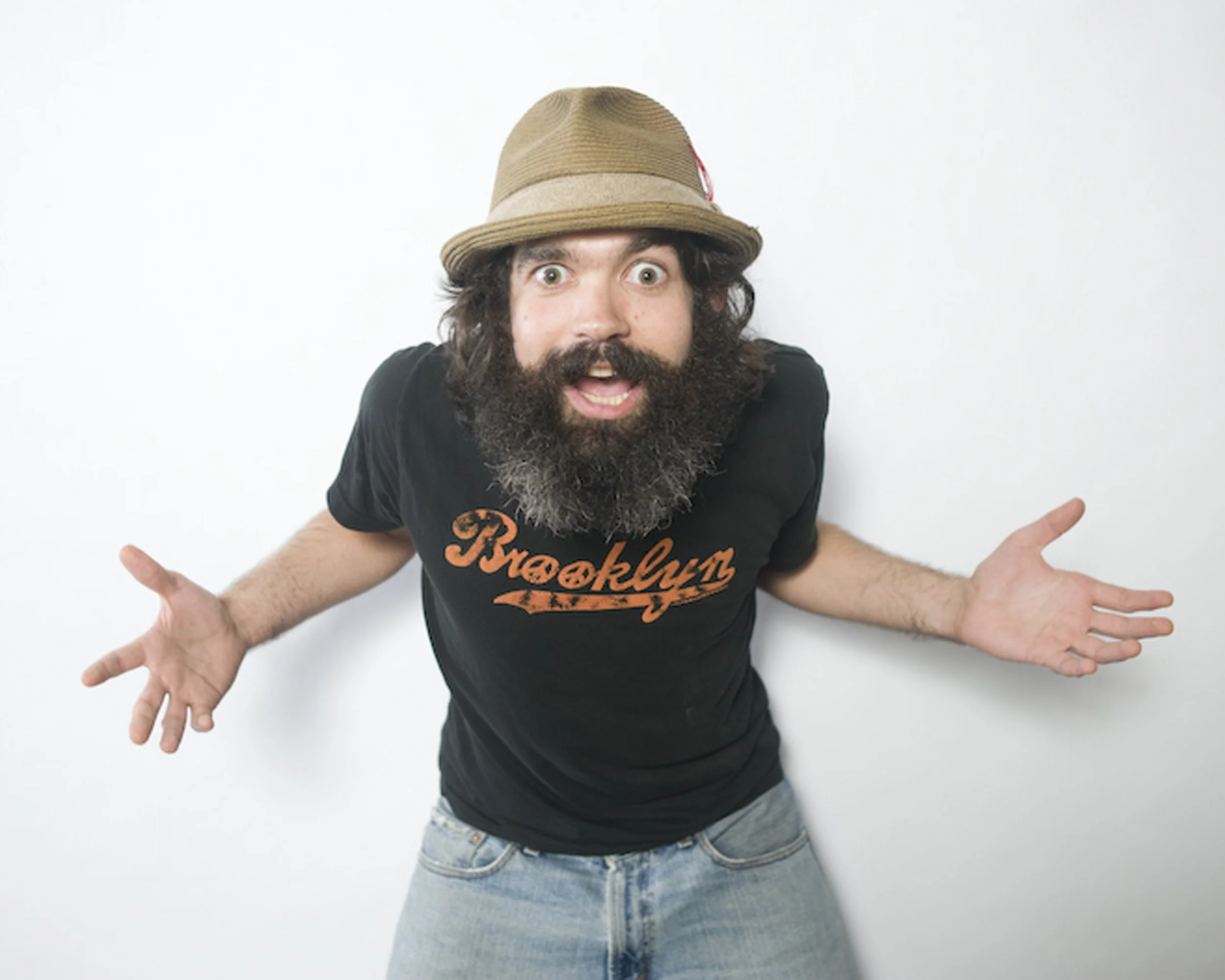 A man with a beard and curly hair wearing a tan hat, black 'Brooklyn' T-shirt, and jeans, standing against a white wall with his arms outstretched and a surprised expression.
