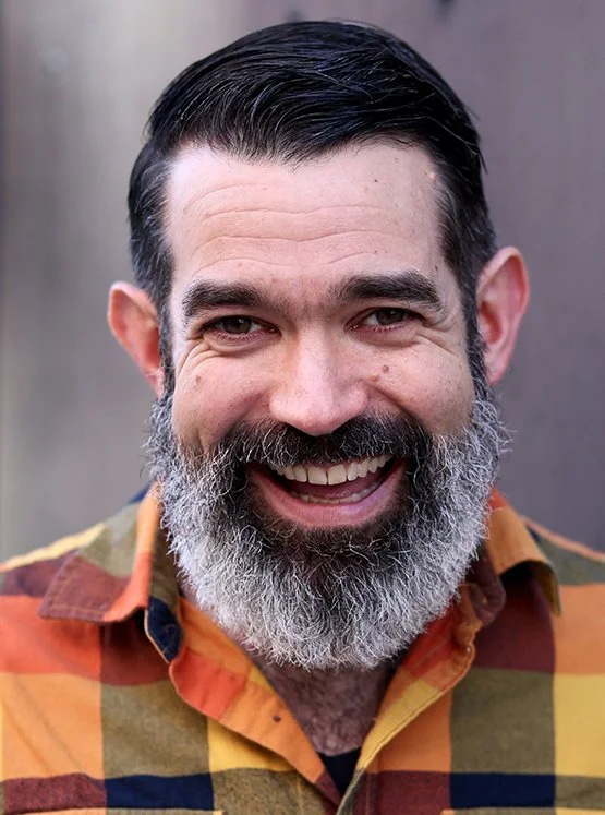 A smiling man with a dark, slicked-back hairstyle and a full gray beard, wearing a colorful plaid shirt, standing outdoors.