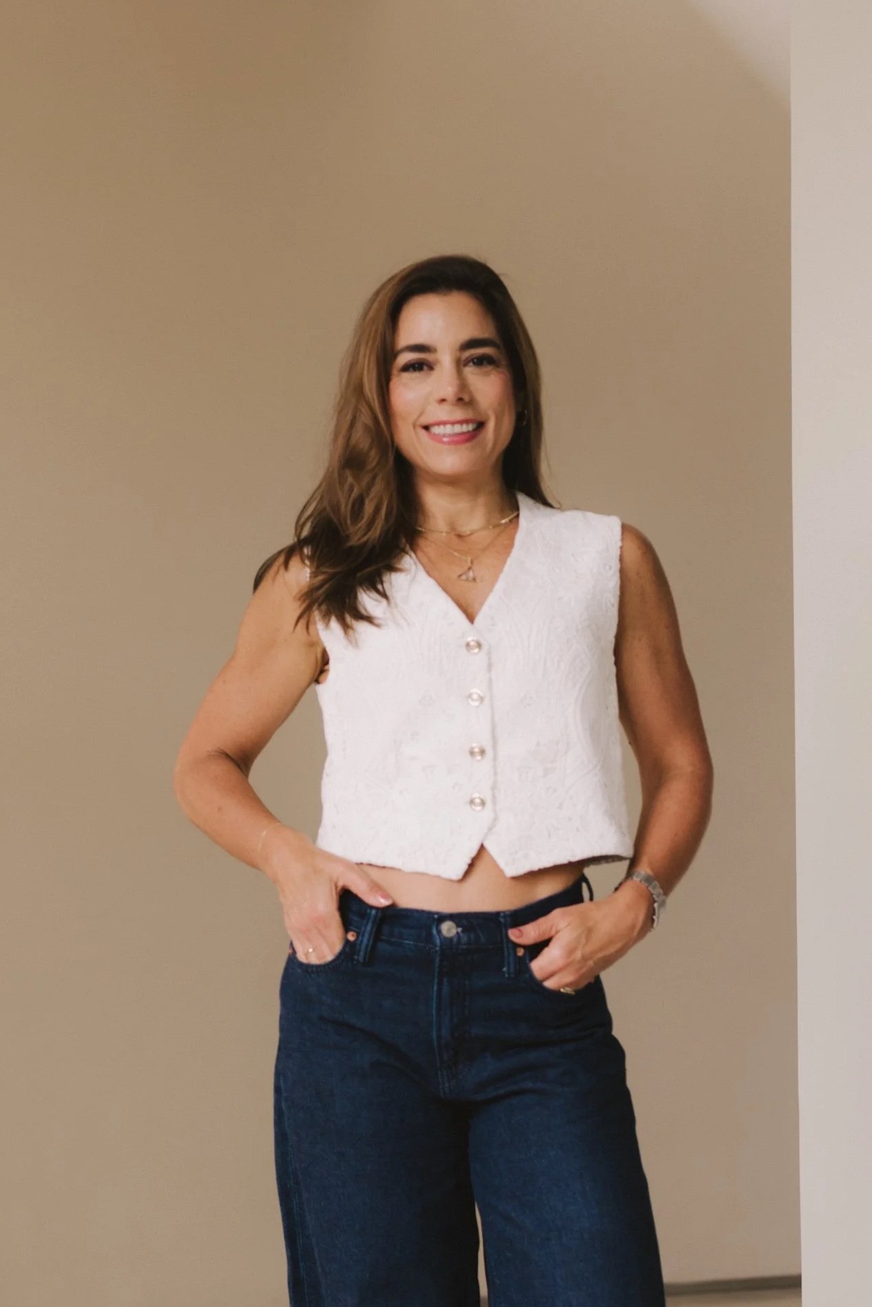 A woman with long brown hair wearing a sleeveless white top and dark blue jeans, standing indoors in front of a beige wall, smiling at the camera.