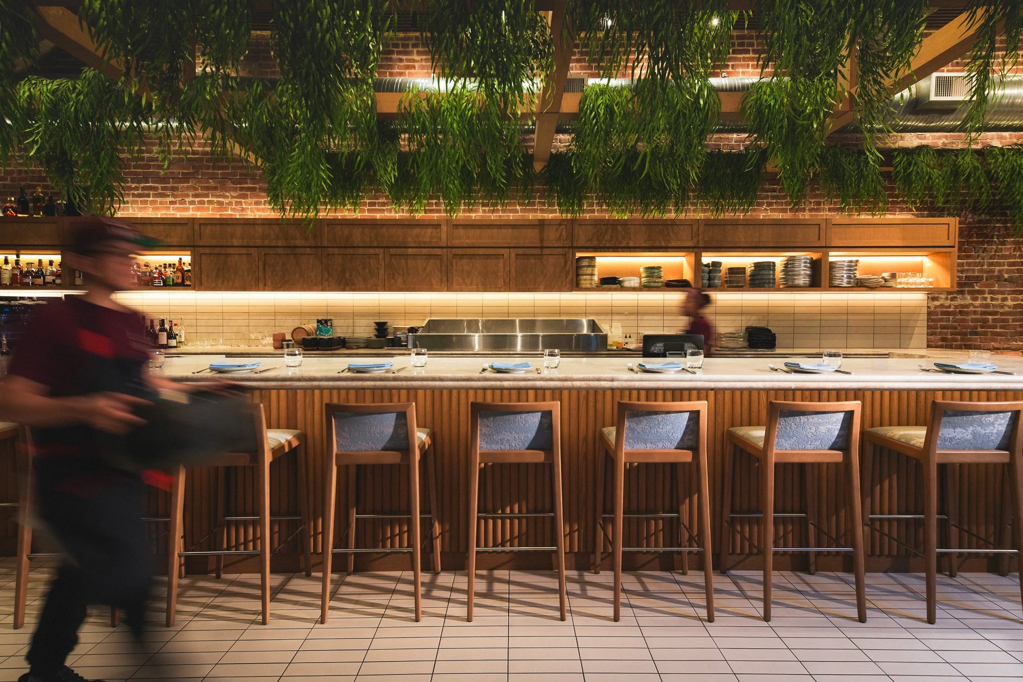 Interior of a restaurant with a long bar counter, wooden bar stools, a brick wall in the background, and hanging green plants overhead. A bartender or server is walking past, slightly blurred.