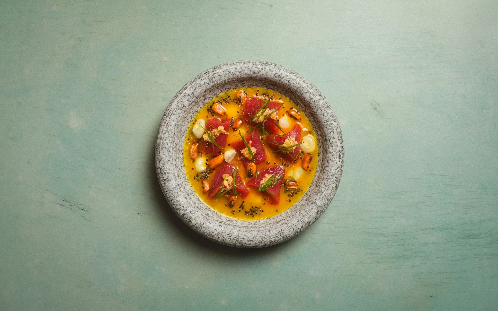 A ceramic bowl with yellow soup, garnished with chunks of raw fish, green herbs, and black seeds, on a light green surface.