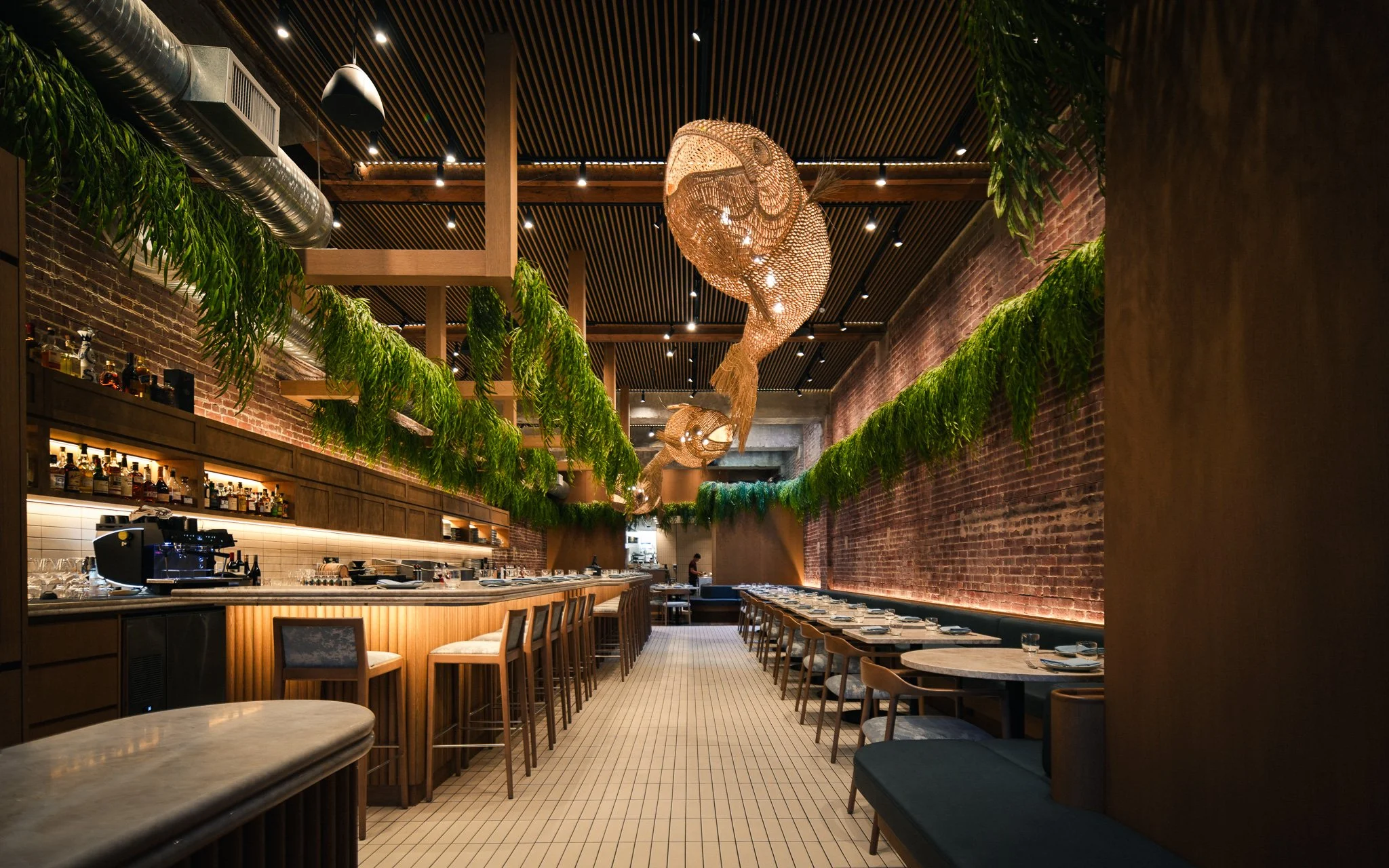 Interior view of UCHU Cevicheria with its exposed brick walls, wooden ceiling beams, and hanging green plants. There is a bar counter with stools on the left and tables with chairs on the right. Decorative woven fish sculptures hang from the ceiling.