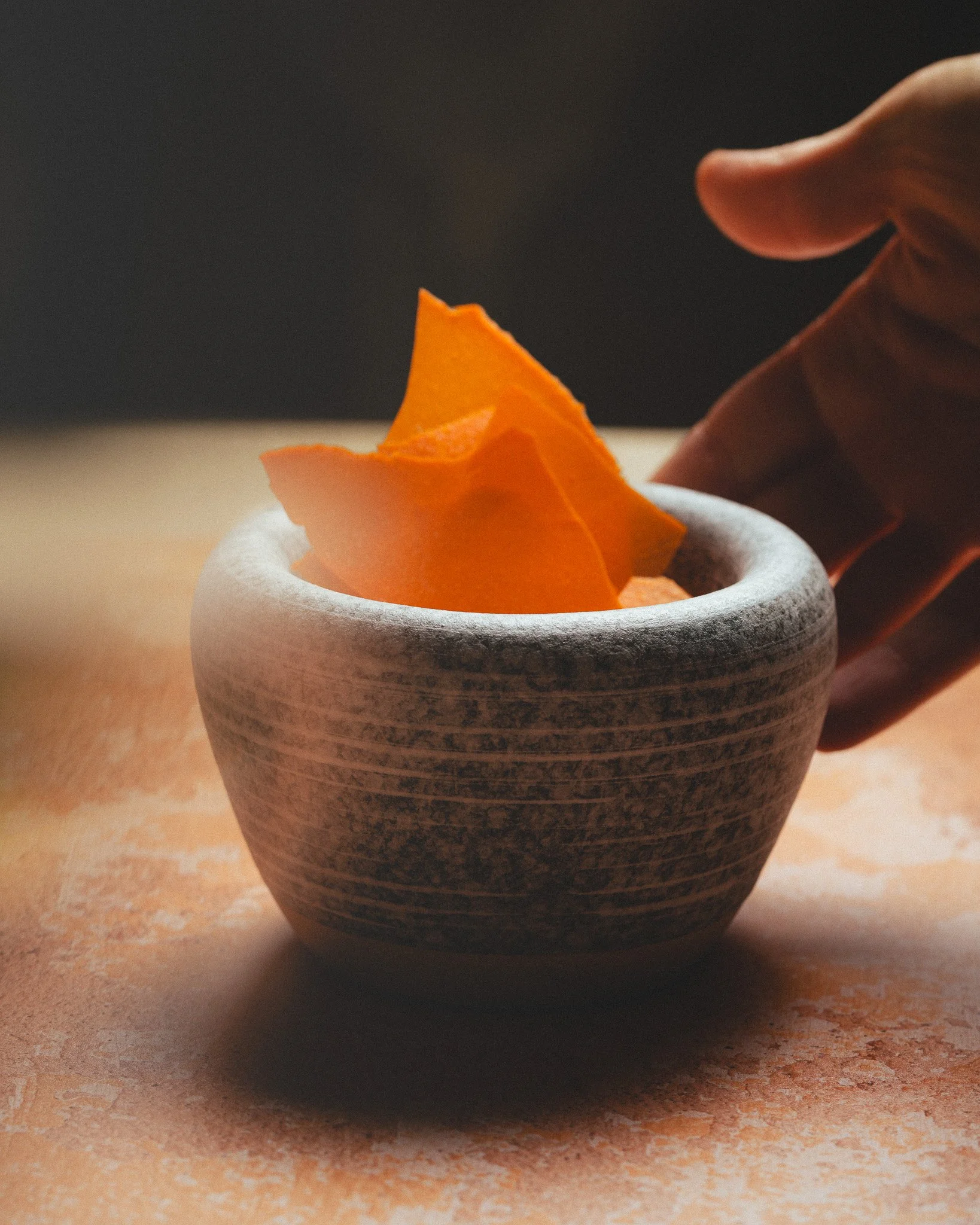 A hand reaching towards a gray ceramic bowl containing bright orange slices, placed on a light-colored surface.