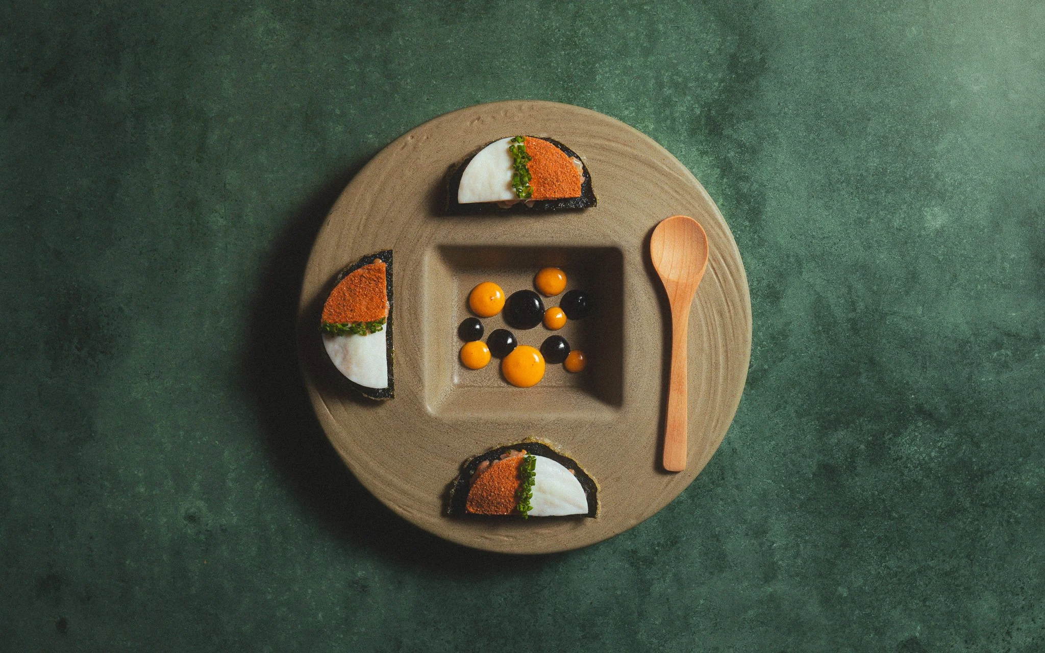 A round plate with a sectioned design, holding three pieces of food with black, orange, white, and green toppings, and a small wooden spoon on a green surface.