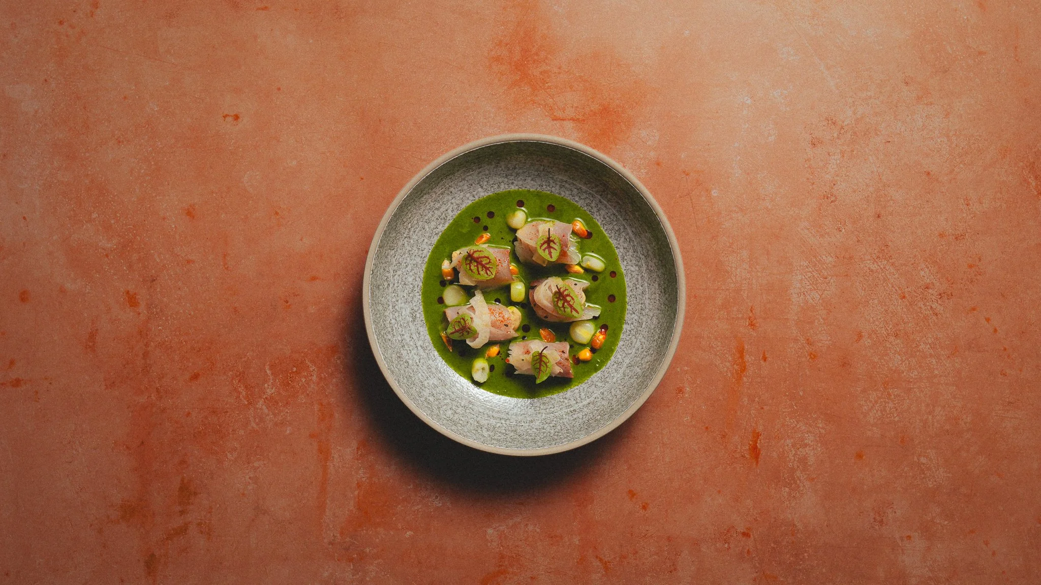 A dish with sliced fish garnished with microgreens, drizzled with green sauce, served in a gray bowl on a textured orange surface.