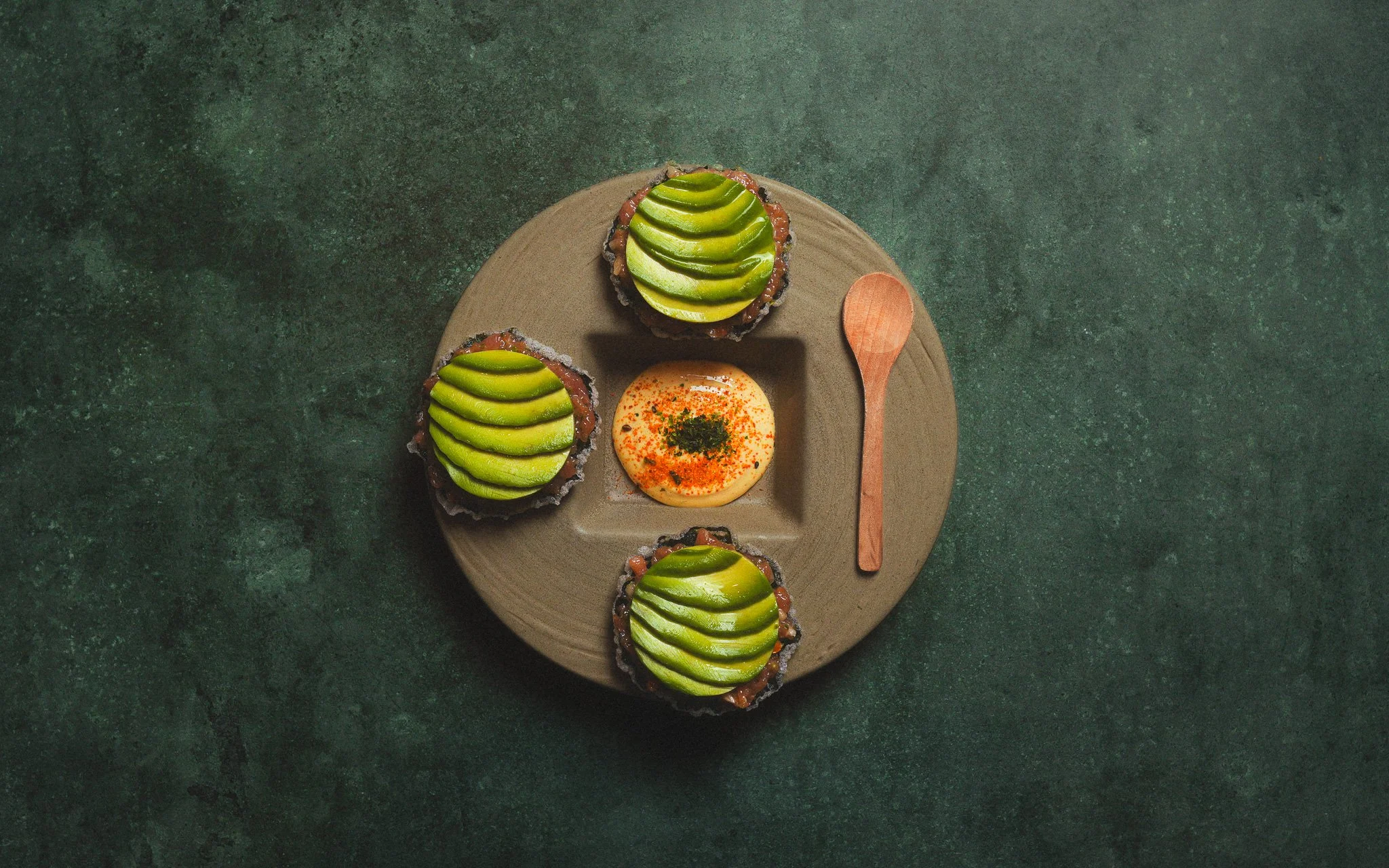 Top view of a round plate with four servings of avocado toast, each topped with sliced avocado, and a small dish of dipping sauce sprinkled with red spices, on a dark green textured surface.