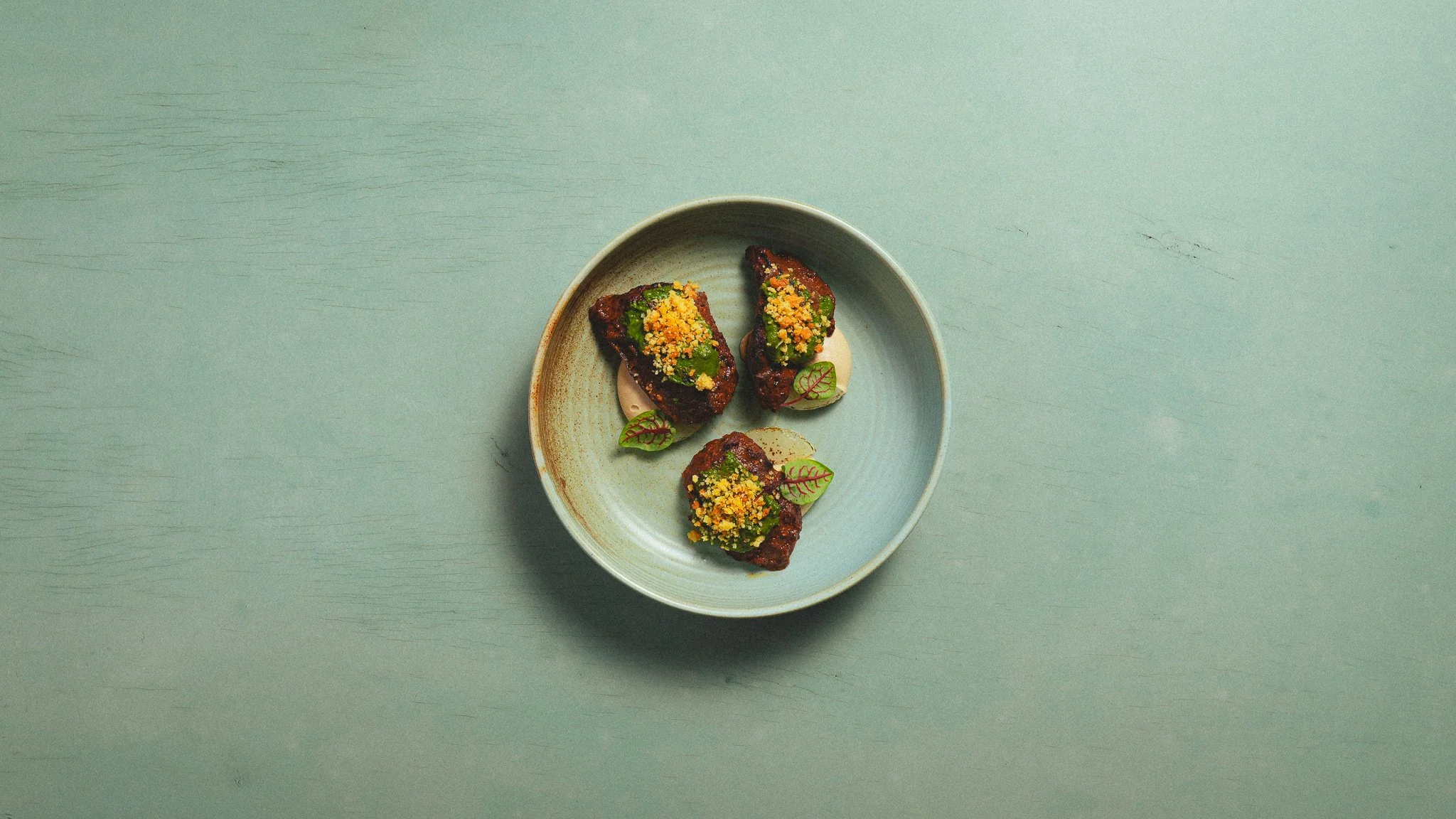 Three pieces of cooked meat garnished with herbs and yellow crumbs on a light blue ceramic plate, placed on a light green wooden surface.