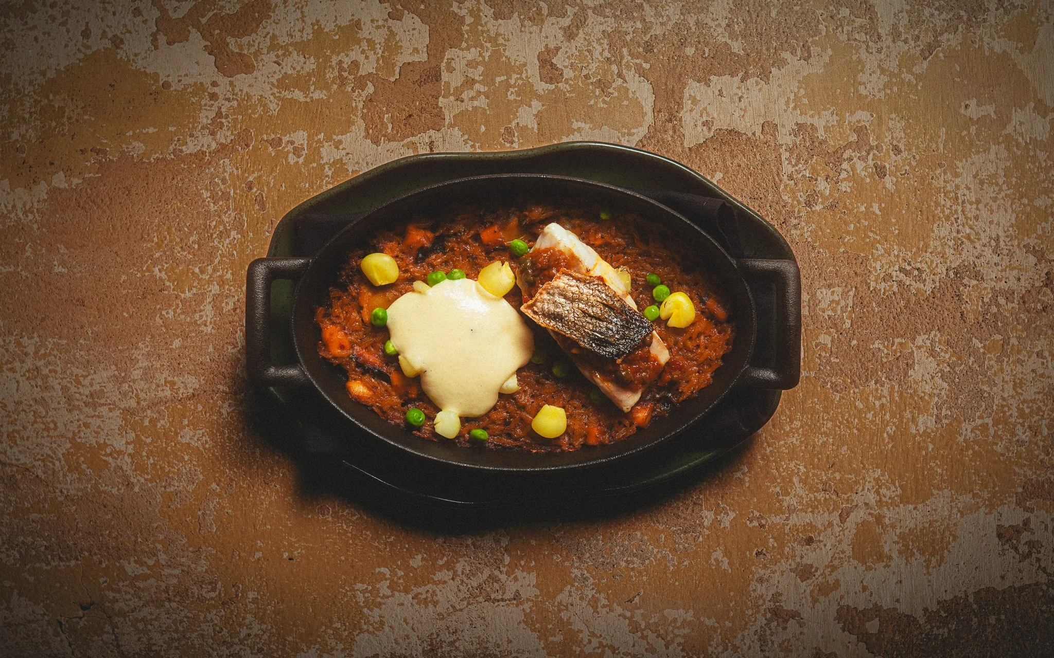 A cast iron skillet with fish topped with melted cheese, served over a tomato-based sauce with peas and corn, on a textured brown surface.