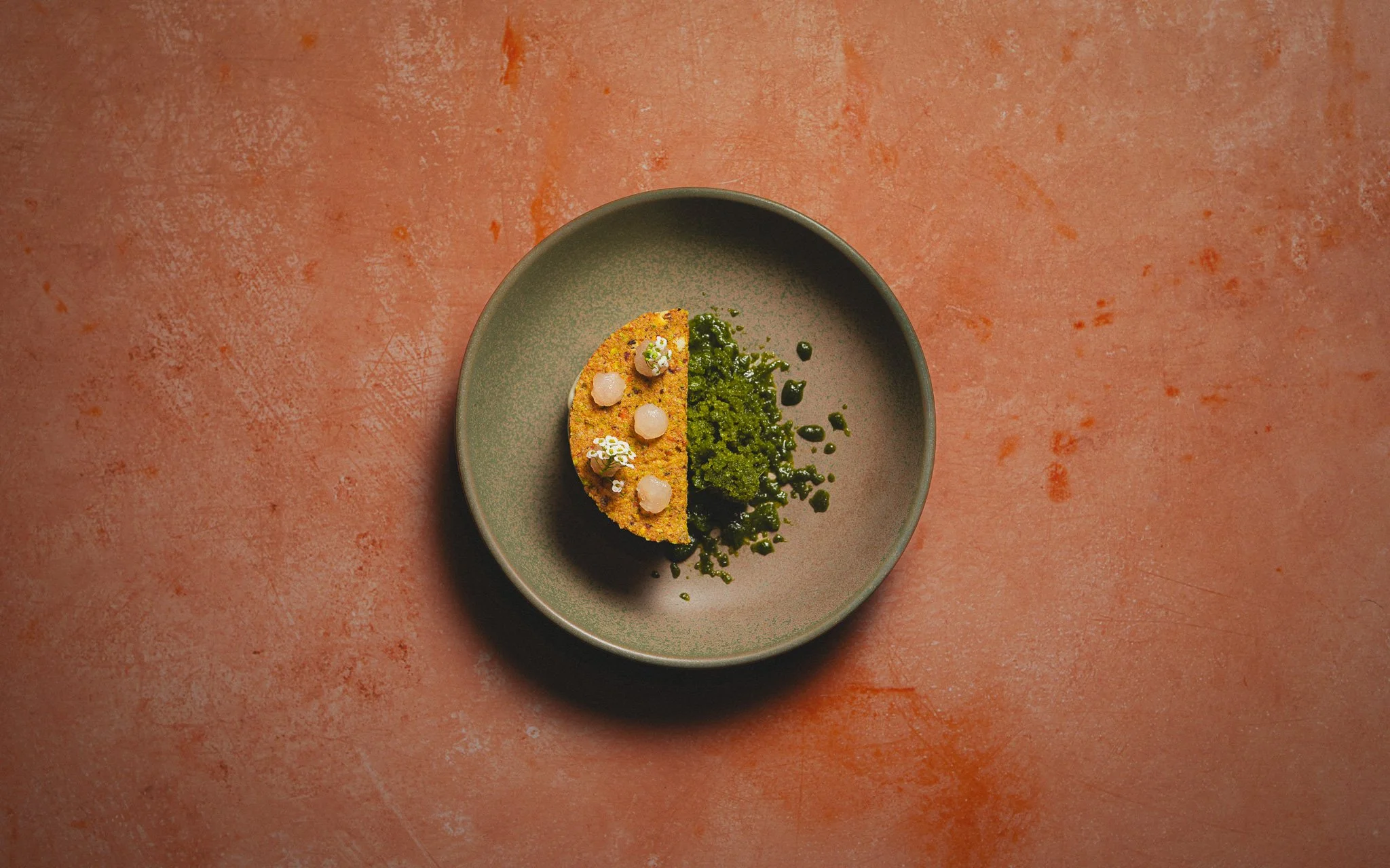 A small, round, gray plate holds a slice of food that appears to be a savory pie or quiche with a golden crust, topped with small white spheres and garnished with edible flowers, alongside green pesto or herb sauce.