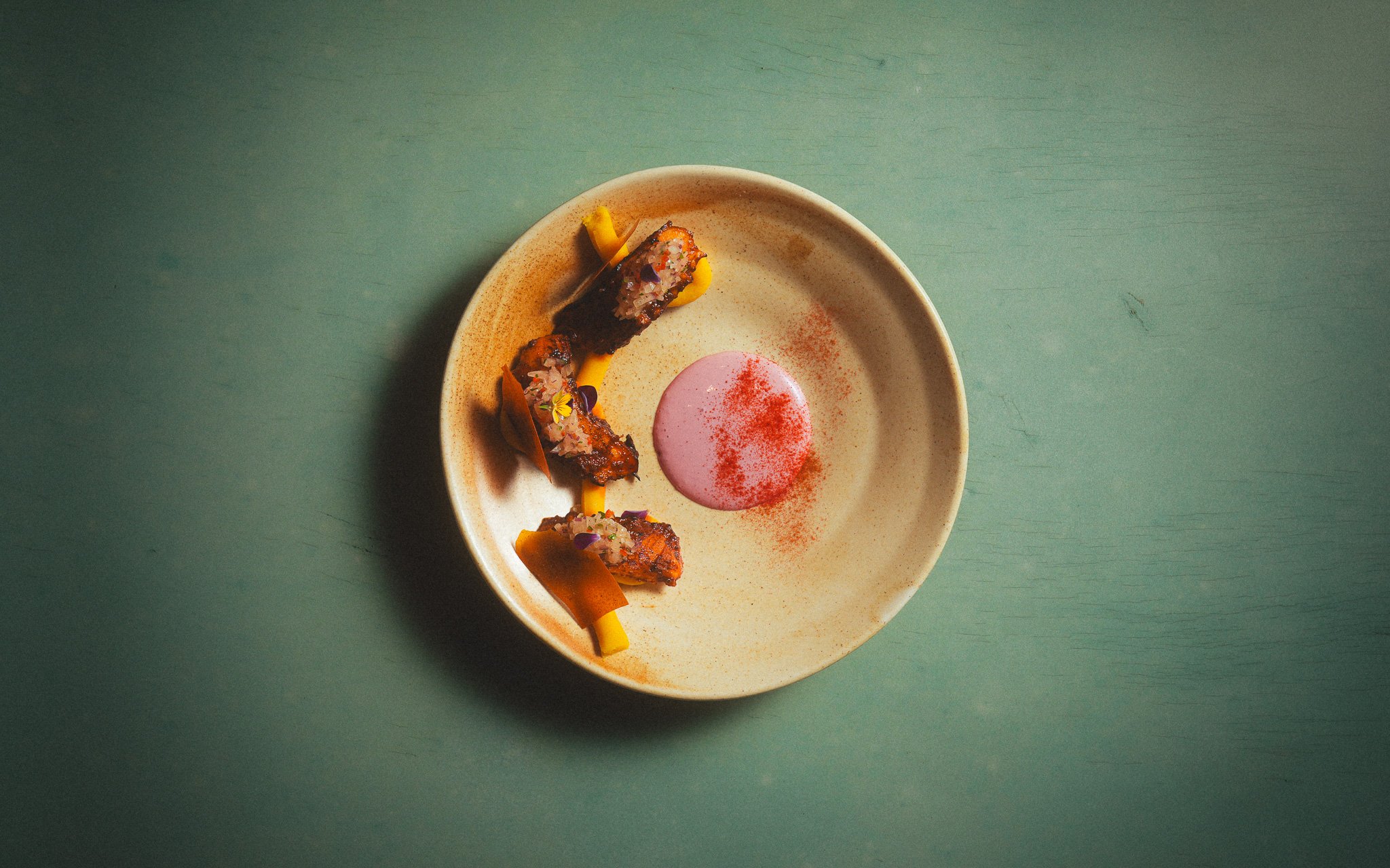 A plate with three pieces of sushi topped with sauce and garnished with edible flowers, accompanied by a pink dipping sauce on a green table.