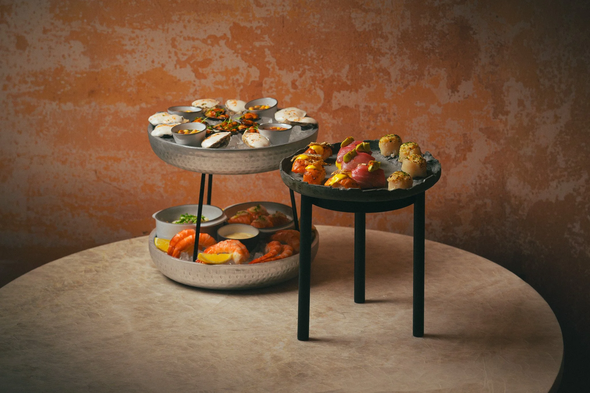 A multi-tiered serving tray with various appetizers, including shrimp and small bowls of sauces, set on a beige table against a textured orange wall.