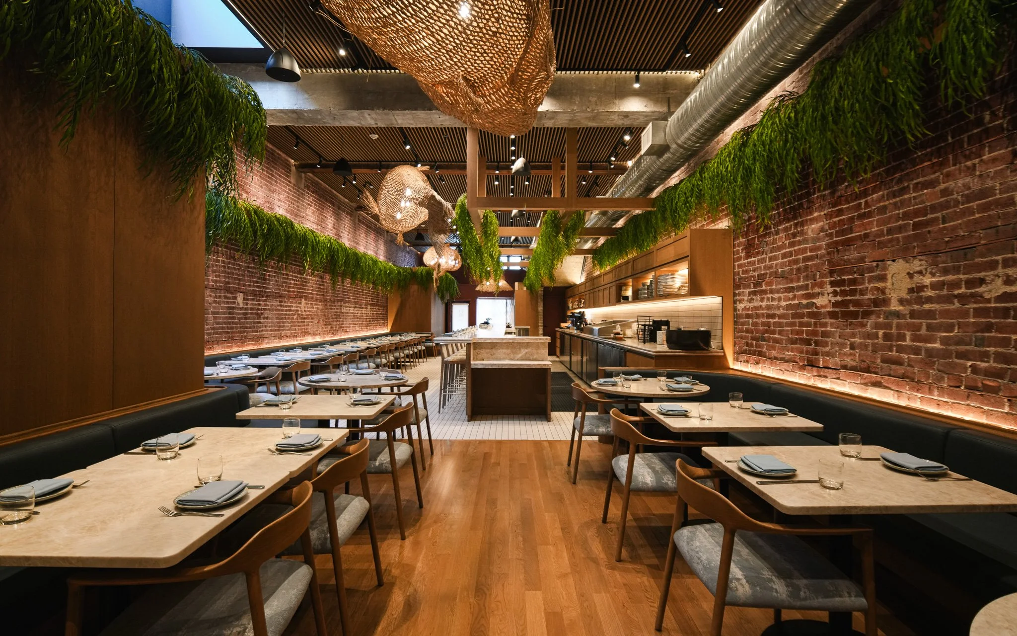 Interior of a modern restaurant with brick walls, green hanging plants, wooden ceiling, and wicker light fixtures, set with tables and chairs.