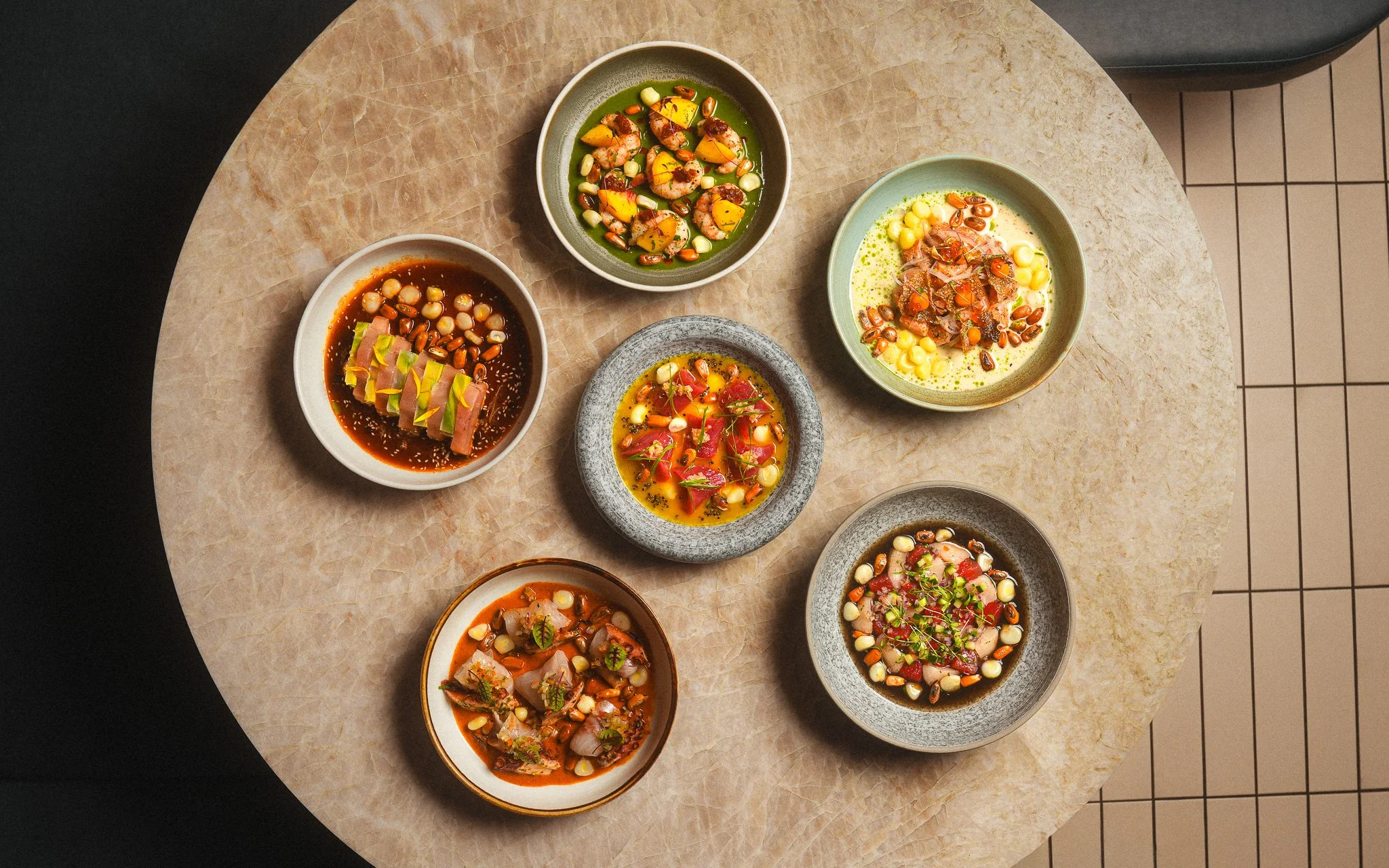 Six different bowls of UCHU's gourmet dishes with various ingredients, including shrimp, vegetables, herbs, and sauces, on a round marble table.