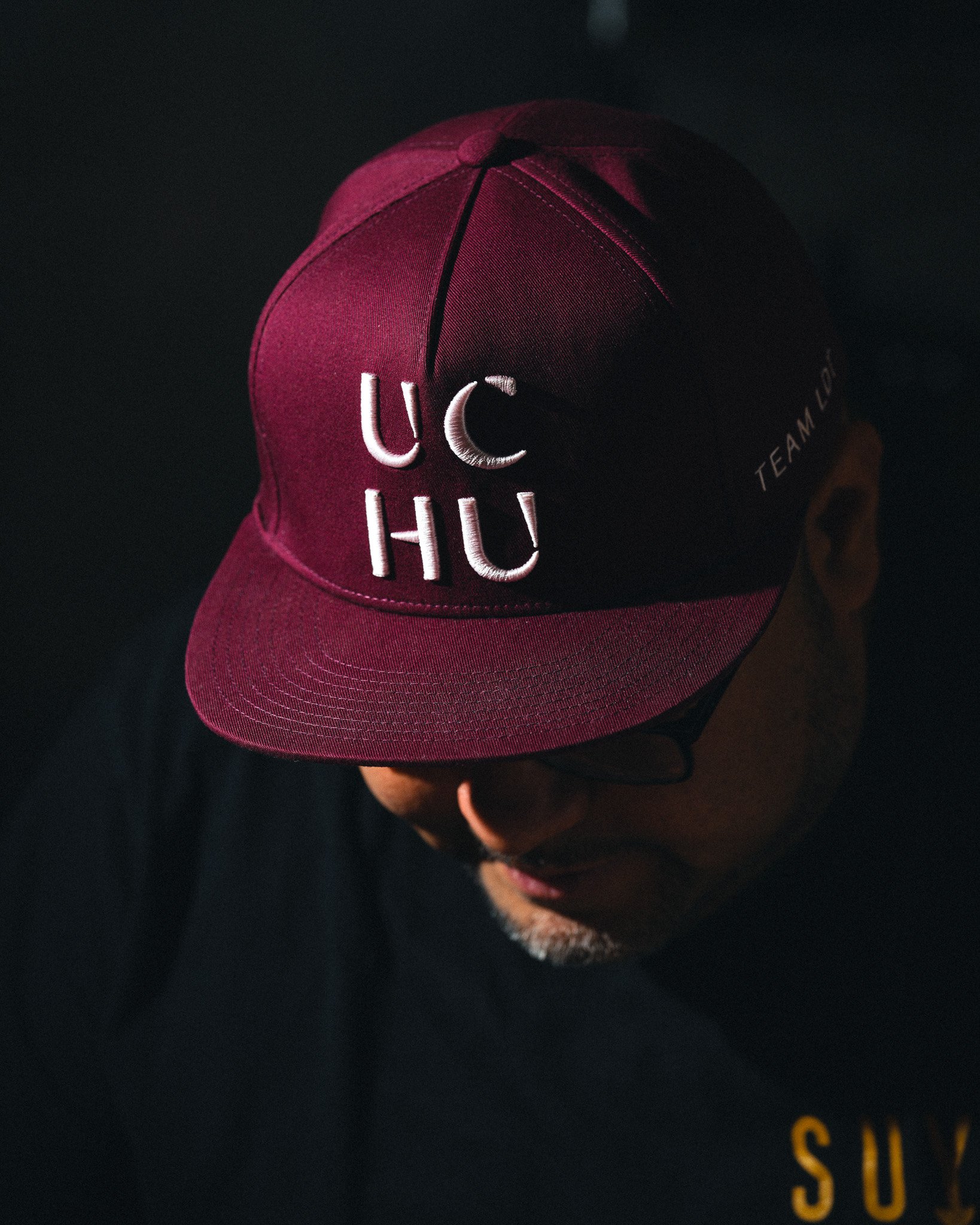 A man wearing a maroon cap with white embroidery that reads 'UCHU' and ‘TEAM’ on the side, with face partially obscured, in low lighting.