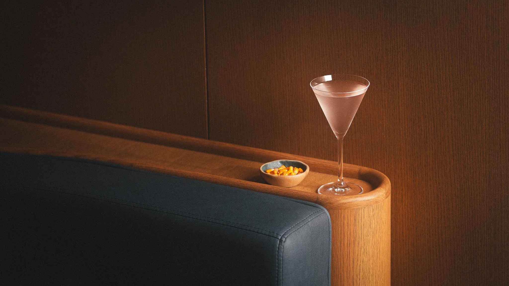 A cocktail glass with a drink on a wooden surface, accompanied by a small bowl of food, against a wooden wall background.