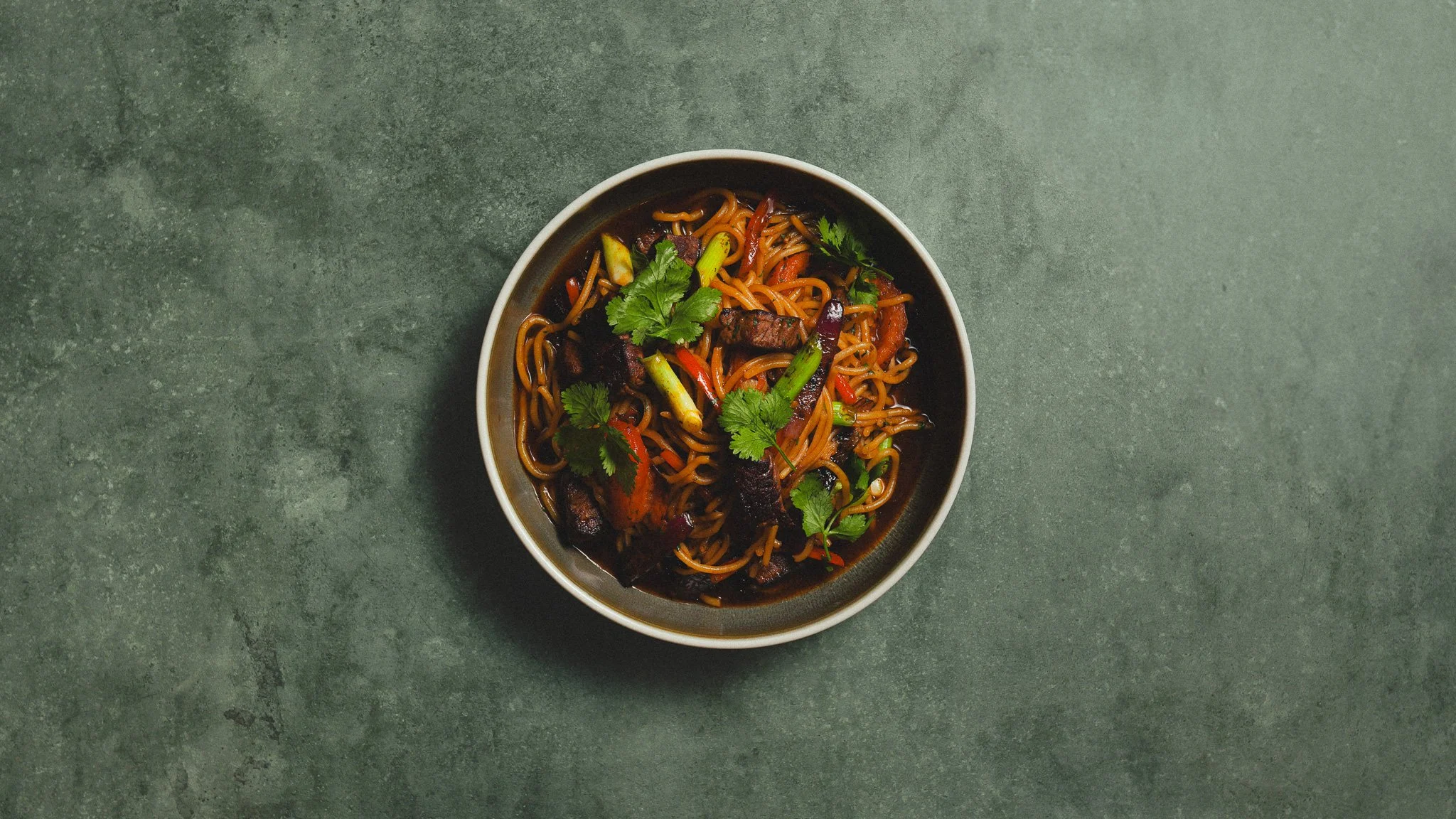 Bowl of stir-fried noodles with vegetables and garnished with cilantro, on a green textured surface.